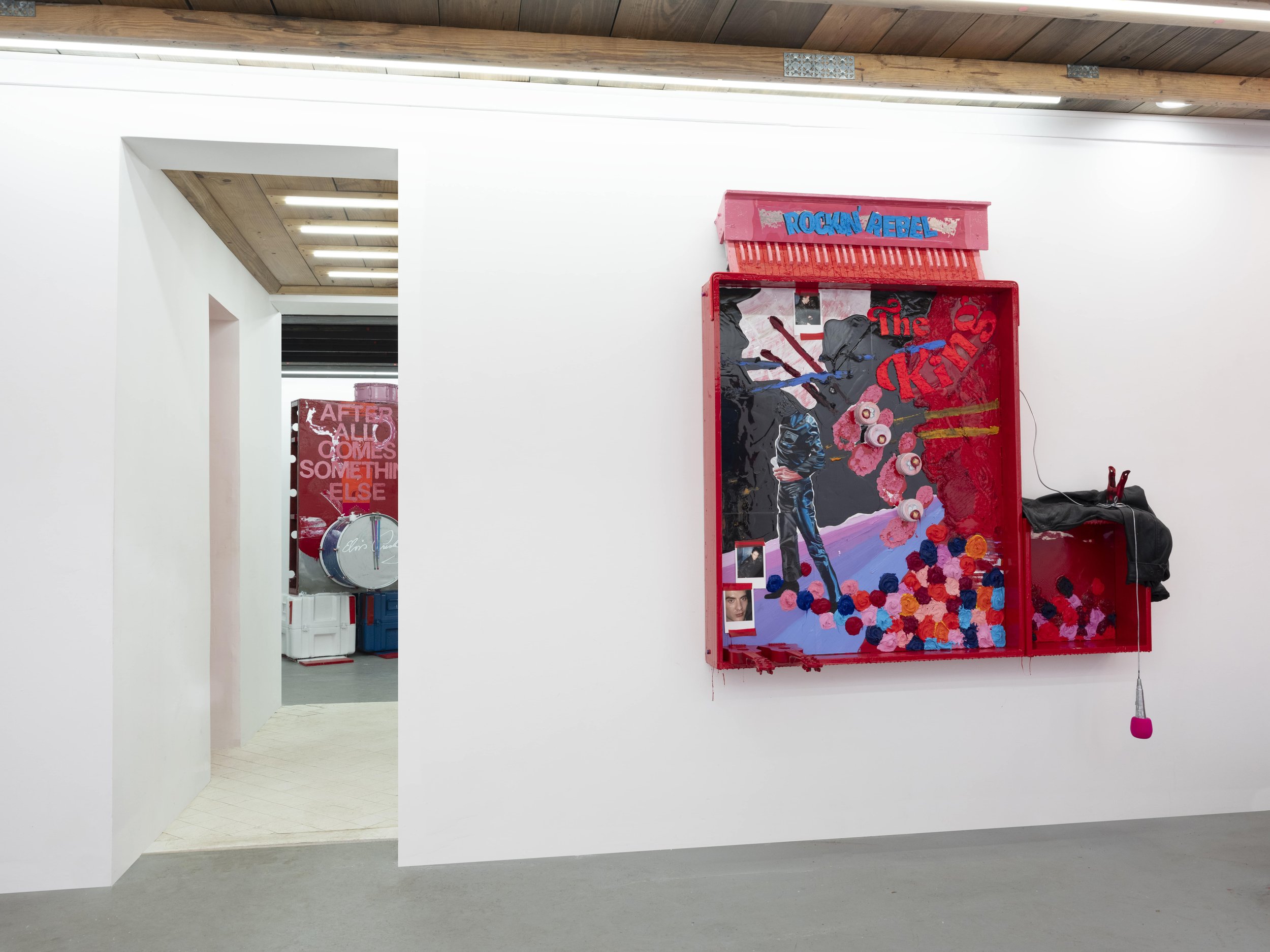 Art installation featuring a large mixed-media piece with a red frame, a figure with a guitar, and colorful pom-poms, with the words 'Rockin' Rebel' and 'The King' in a gallery with white walls and a wooden ceiling.