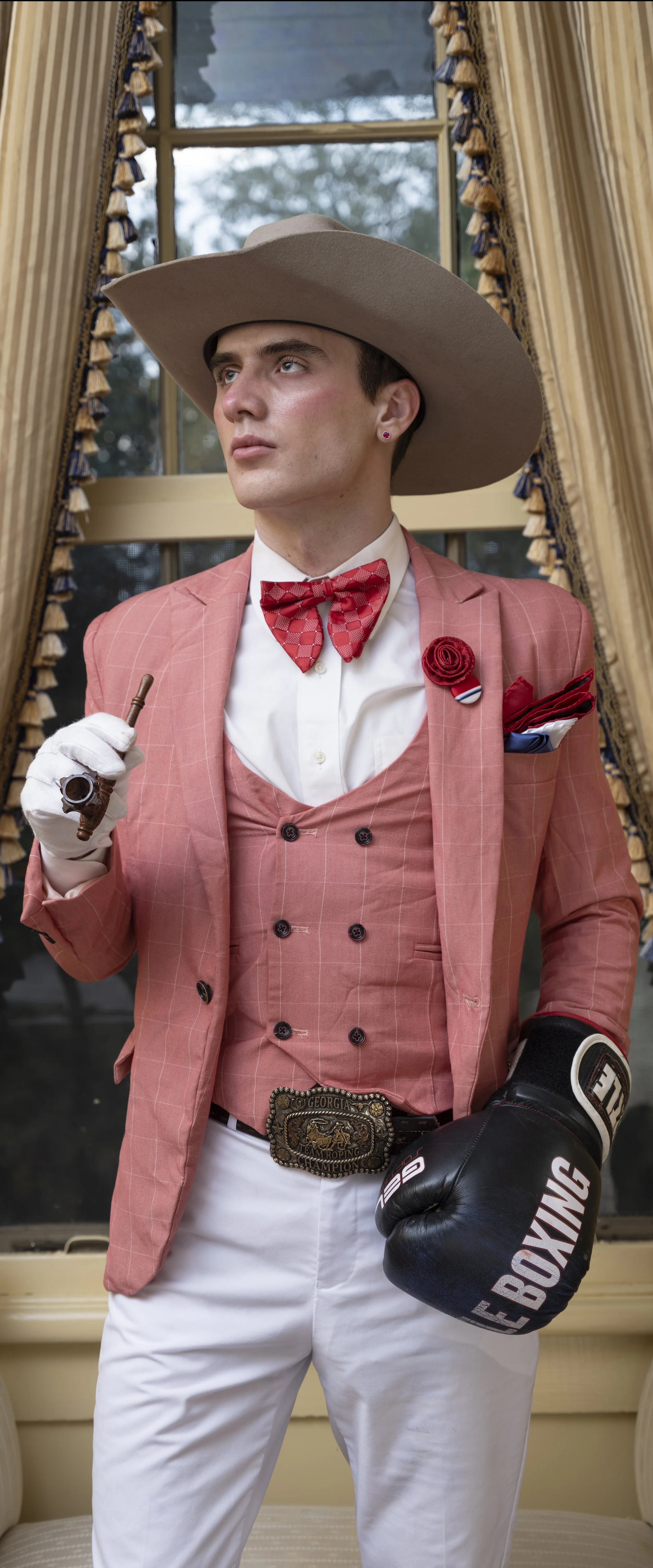 Young man in vintage boxing attire with a large beige wide-brimmed hat, red checkered bow tie, pink blazer, white shirt, pink vest, white pants, and boxing glove, holding a cigar. He is standing in front of a window with gold curtains.
