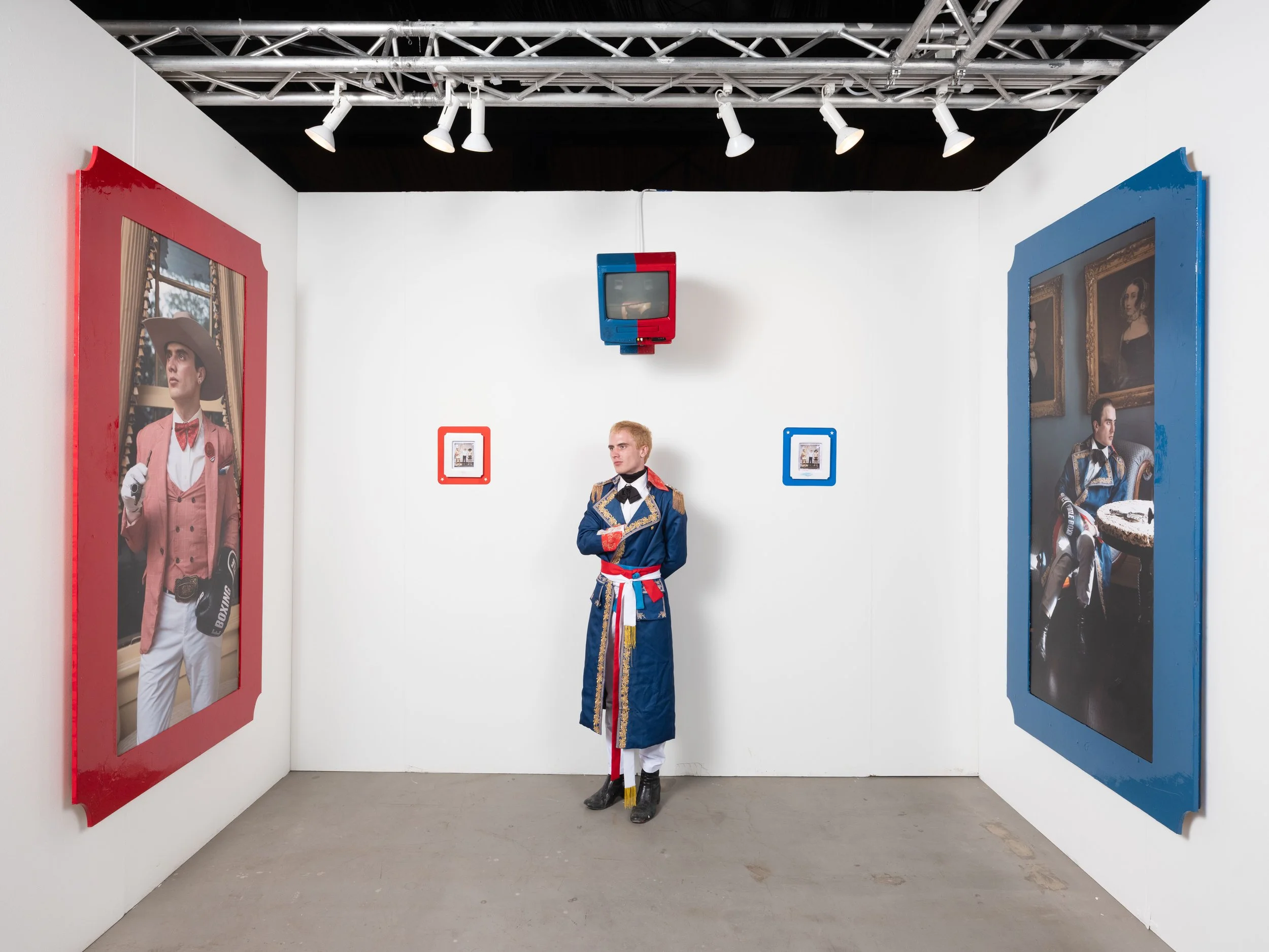 An art gallery with white walls and a gray floor, featuring large framed photographs on both sides. A person dressed in a colorful, ornate costume stands in the center, looking to the left. There are small framed pictures on the wall behind and to the sides of the person. A small red, blue, and black TV is hanging from the ceiling above, and a metal truss with white spotlights is also overhead.