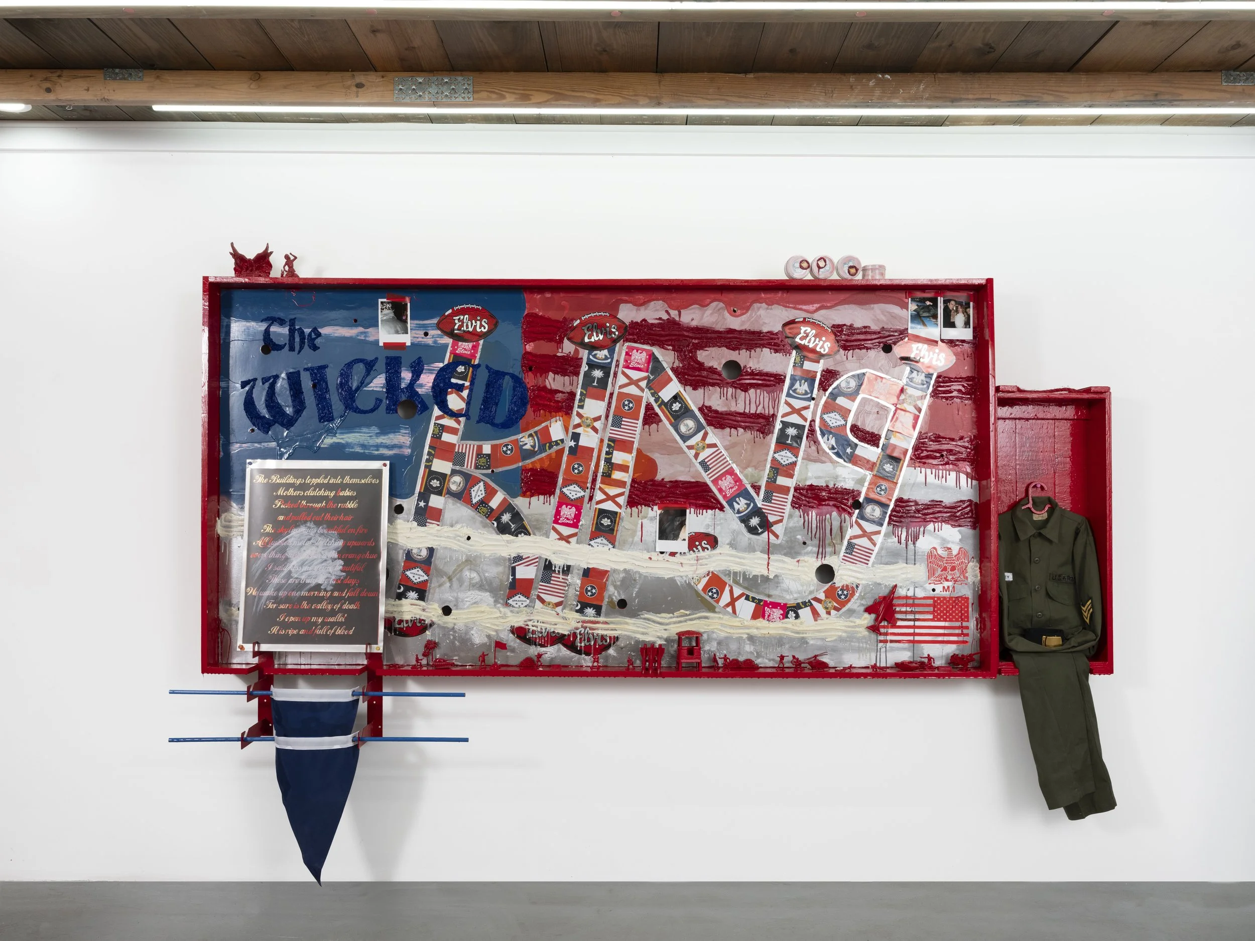 A large American flag-themed art installation with the words 'The Wicked' and 'LOVE' displayed. It features various patches and images, a military jacket hanging on the right side, a blue and white umbrella on the left, and several small photos and decorations, all mounted on a white wall.