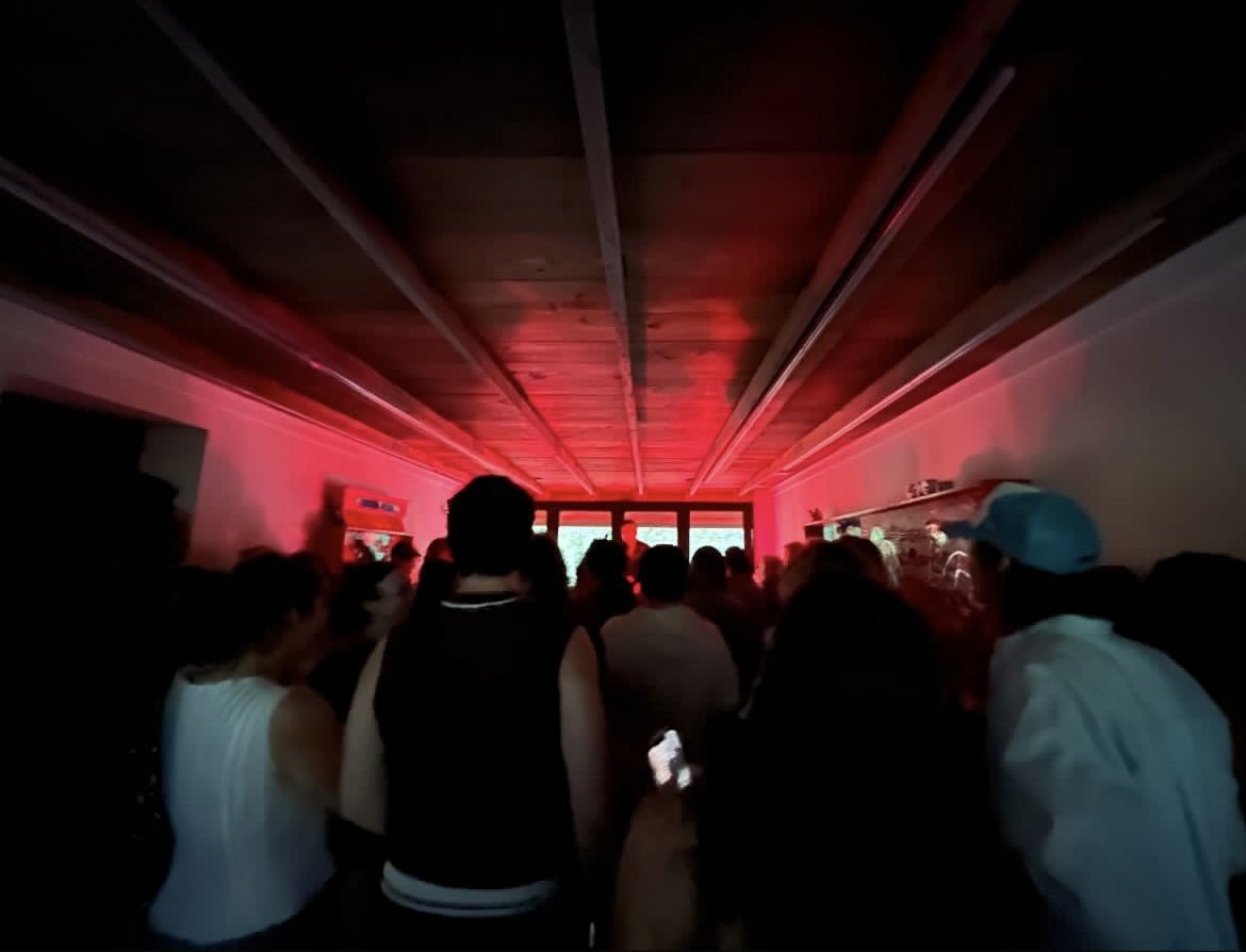 A crowded indoor room with people watching a large screen at the front, illuminated by red lighting, with some individuals holding phones and wearing casual clothing.
