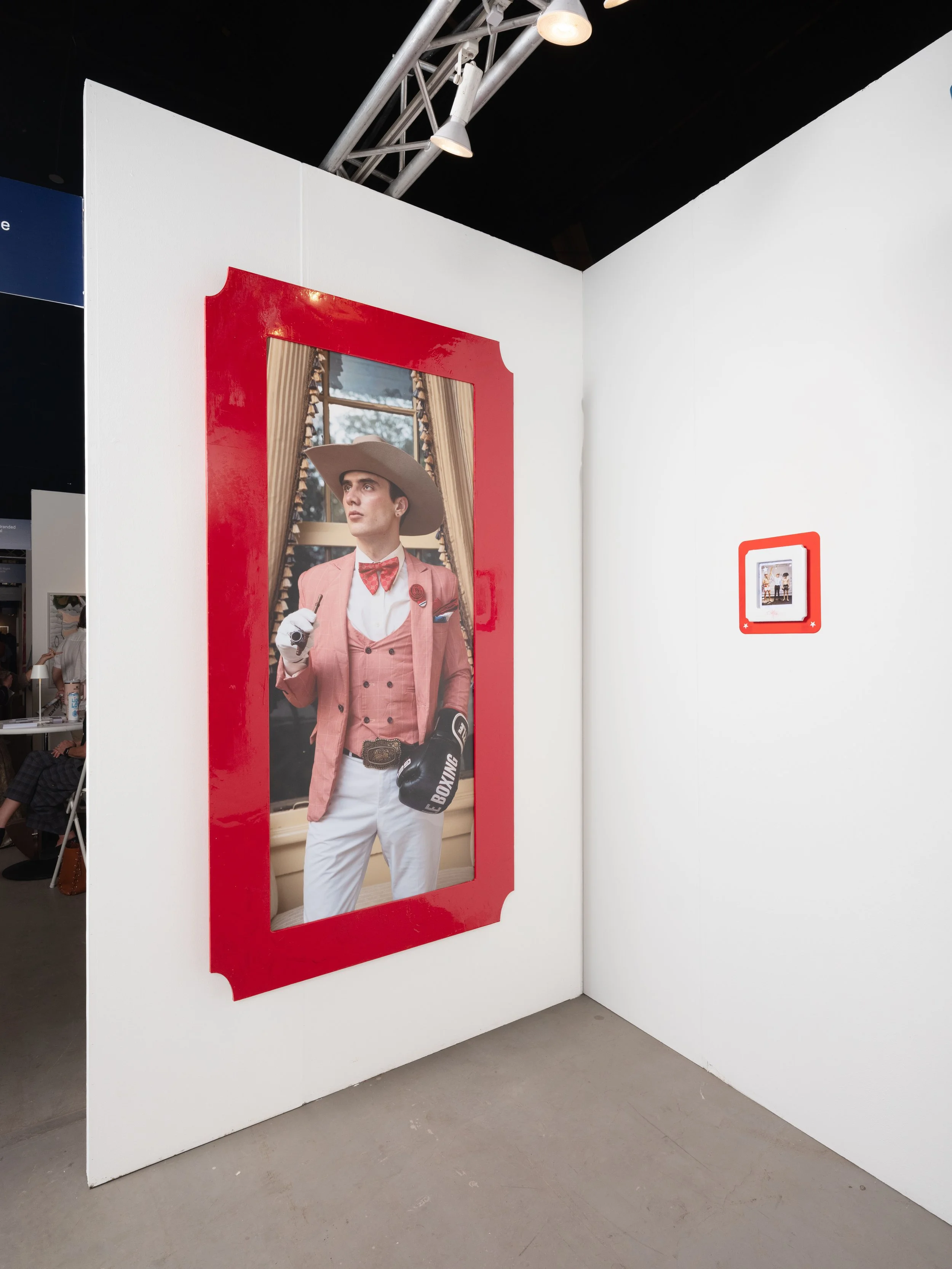 A life-sized cutout of a young man dressed in a pink tuxedo with a cowboy hat, standing in front of a mirror with gold curtains and window.