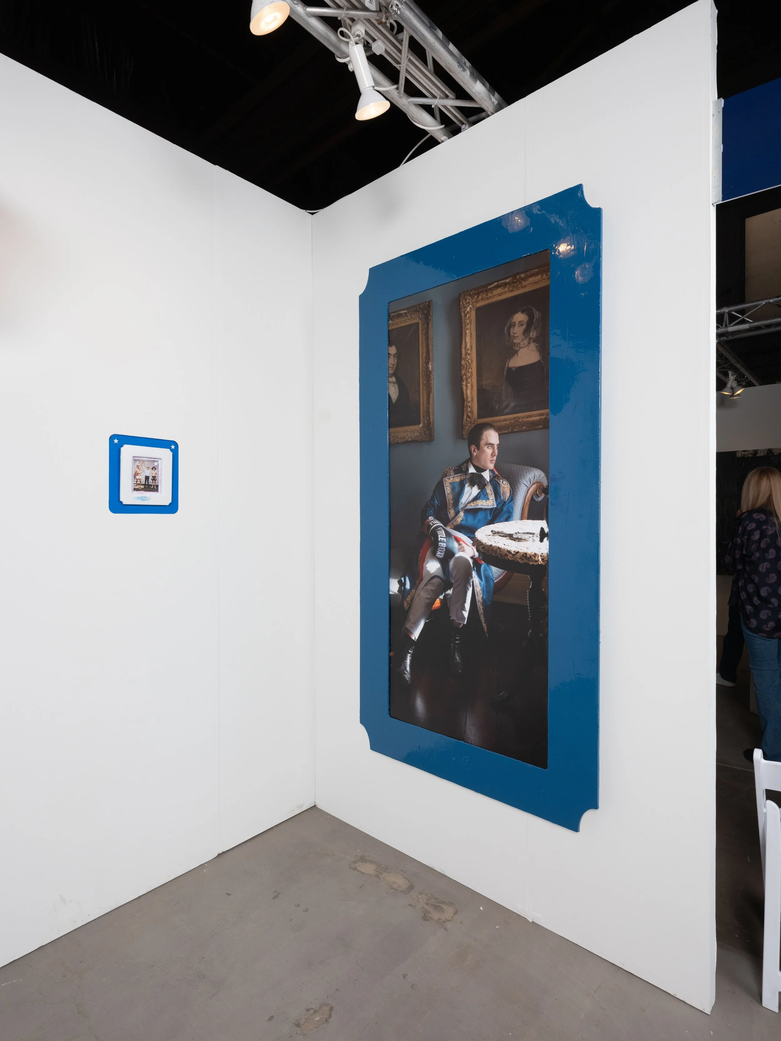 Large portrait of a man dressed in a formal racing suit sitting in a vintage-style room with framed portraits on the wall behind him, framed with a blue border, displayed at an art gallery or exhibition.