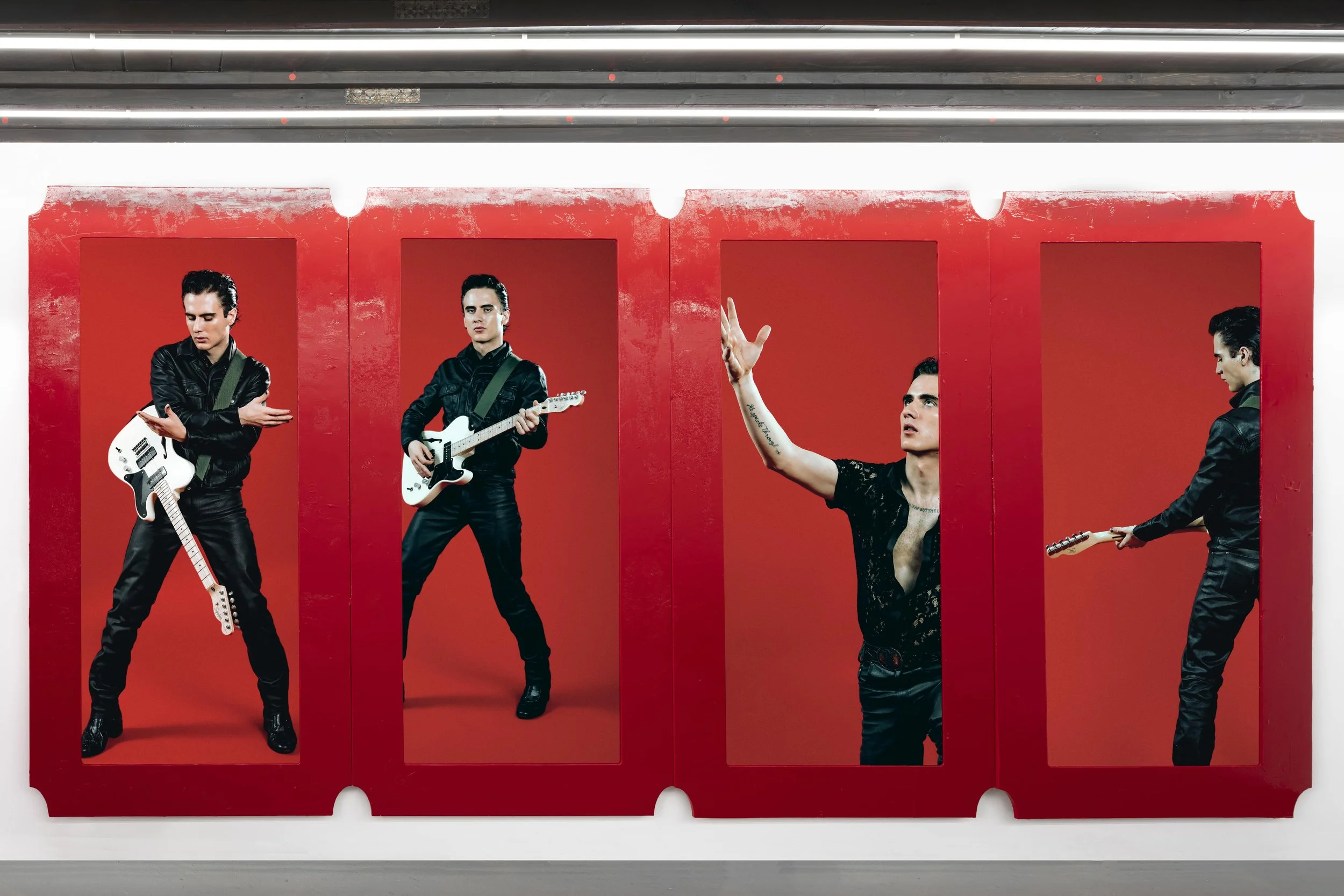 Four life-size posters of a male musician with black hair, dressed in black, with guitars, set in red corners on a white wall, with the third poster showing the musician gesturing with his hand.