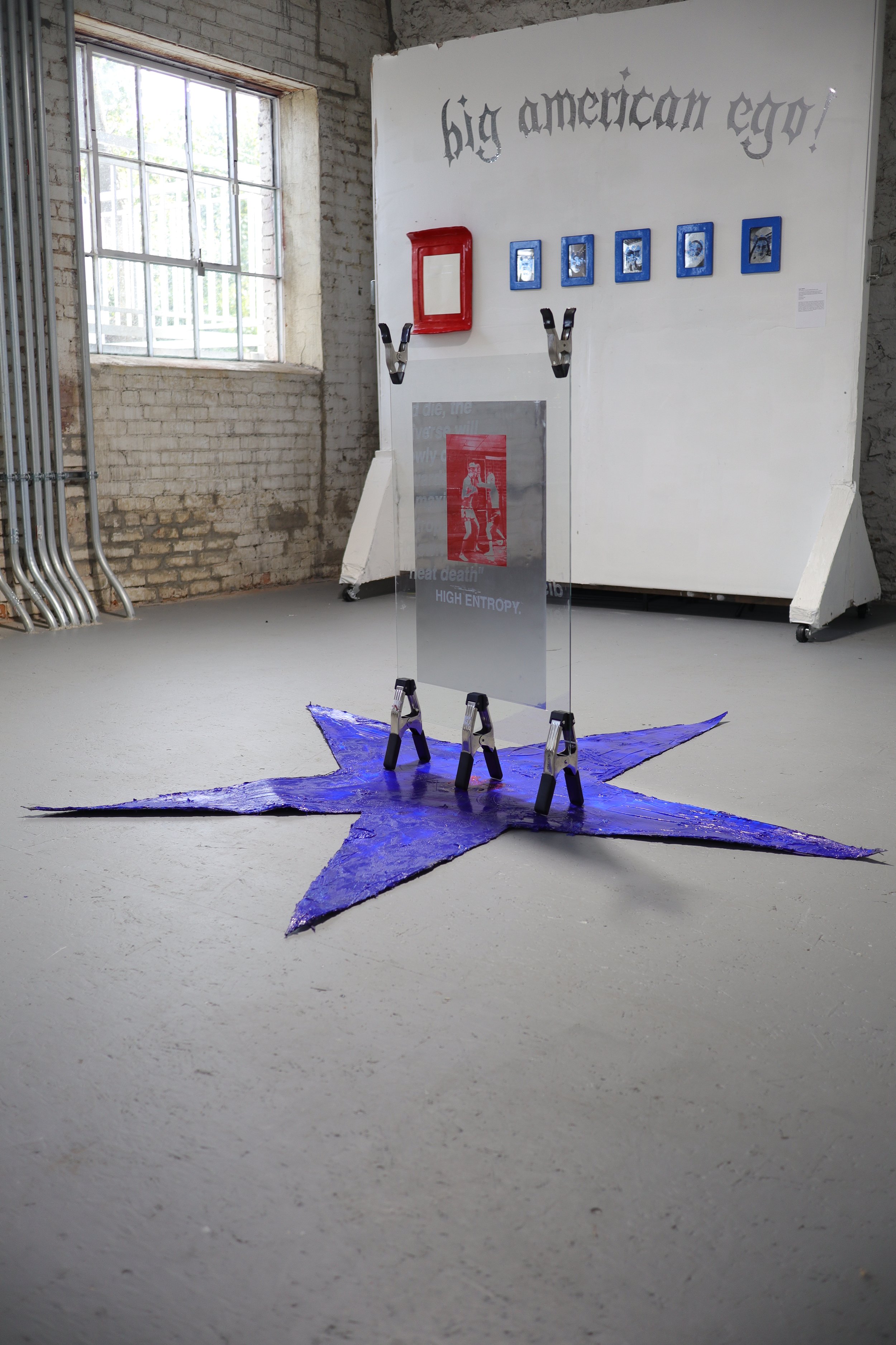 An art installation featuring a large purple star-shaped carpet with four black clamp-like objects holding a transparent panel with red and black text and images, in front of a white wall with the phrase 'big american ego!' written in black, gothic-style lettering, and framed photos above.