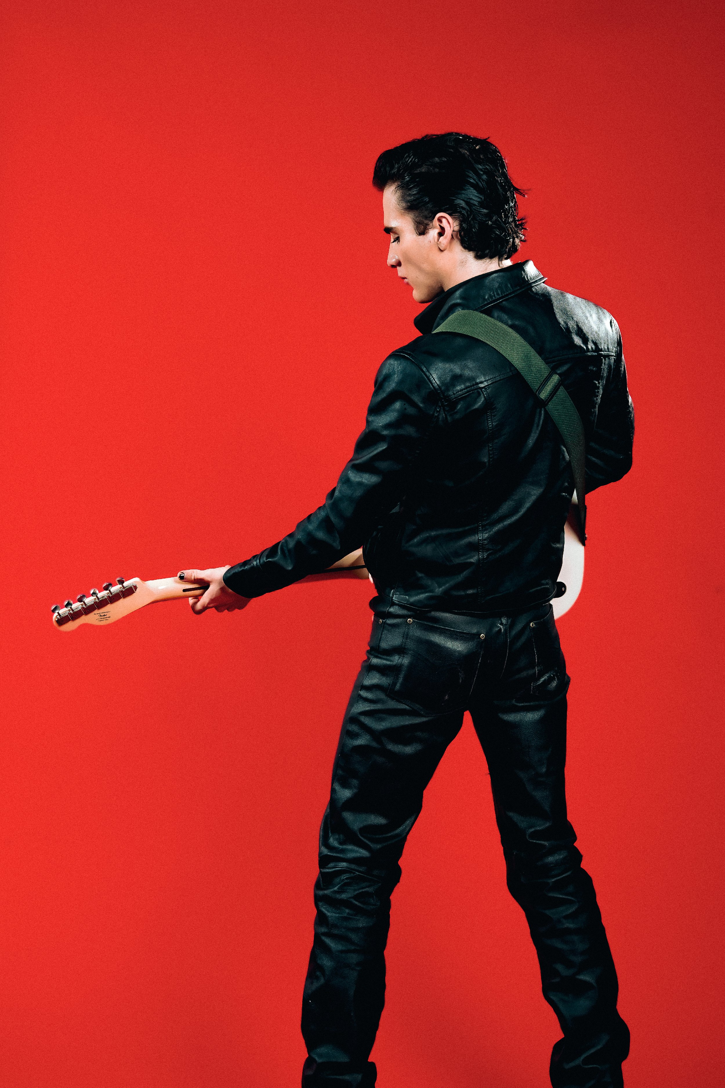 A man with dark hair, wearing a black leather jacket and black jeans, is holding a white electric guitar against a red background.