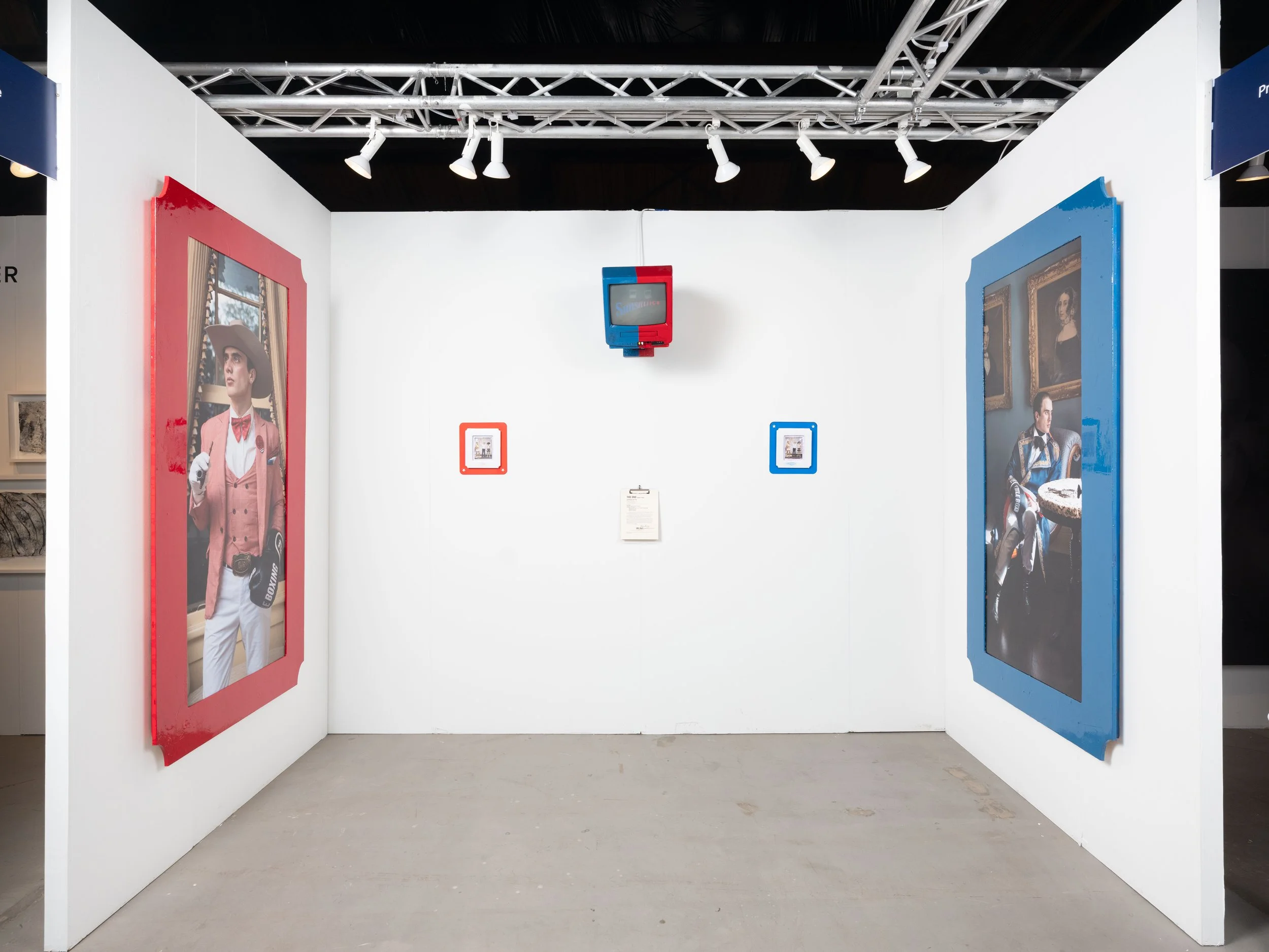 An art exhibit with two large framed photographs on white walls, one on each side. The left photo shows a woman dressed as a golfer or tennis player with a hat and camera, and the right photo features a man in a racing suit sitting in a room with framed paintings. A small screen hangs from the ceiling, and two smaller framed images are on the back wall, one in red and the other in blue. Track lighting illuminates the display.