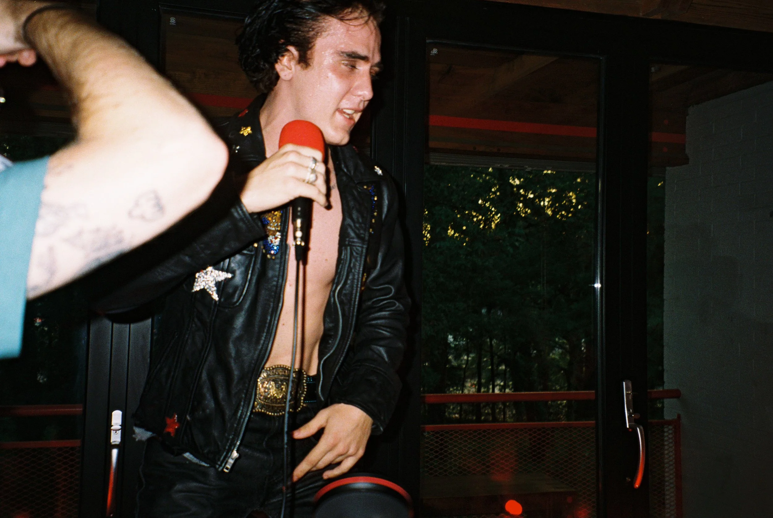 A man performing karaoke or singing into a microphone, wearing a black leather jacket with shiny embellishments, no shirt underneath, and a large belt buckle.