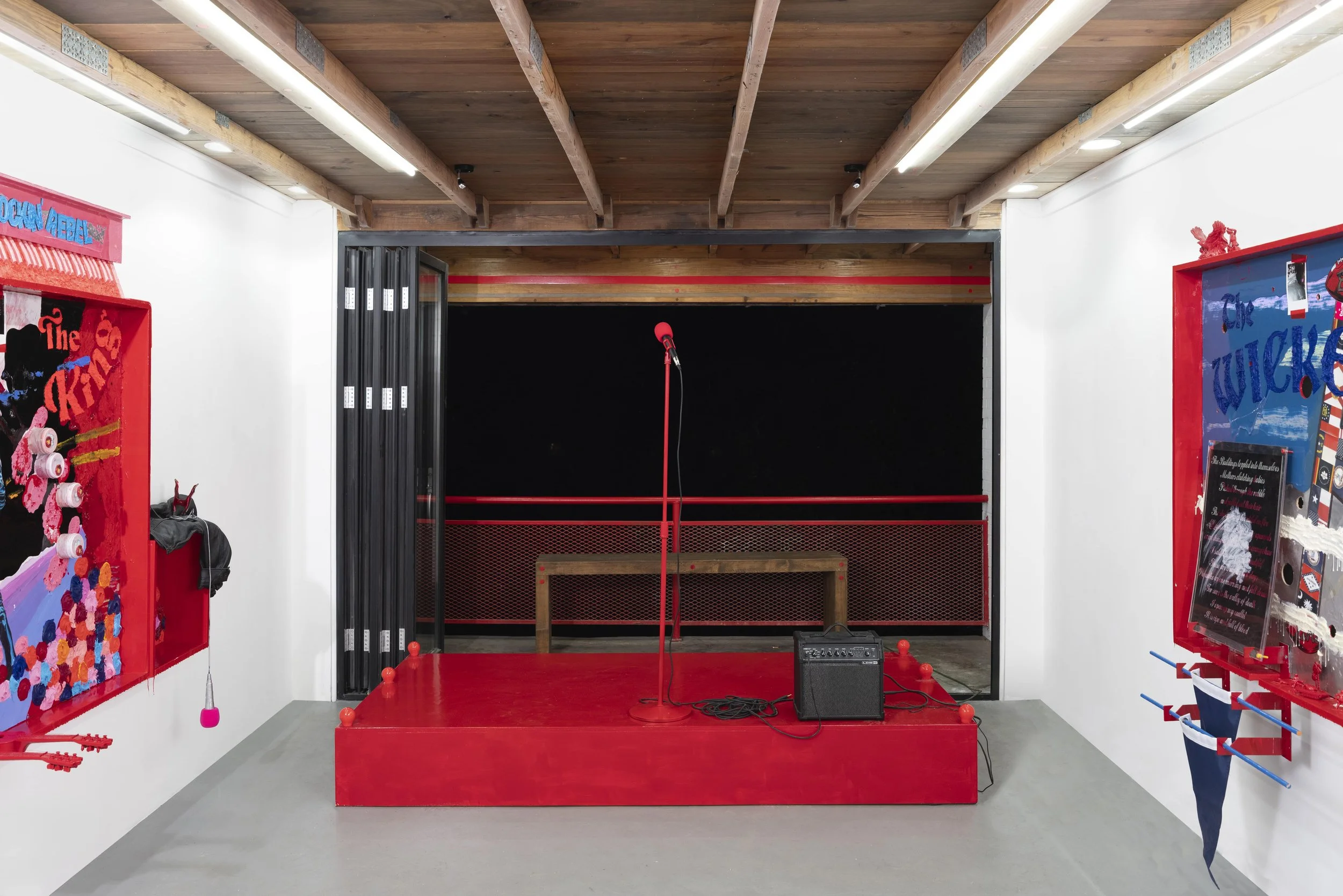 Empty red stage with microphone, black speaker, and red railing, in front of a black background, with colorful wall art on white walls.