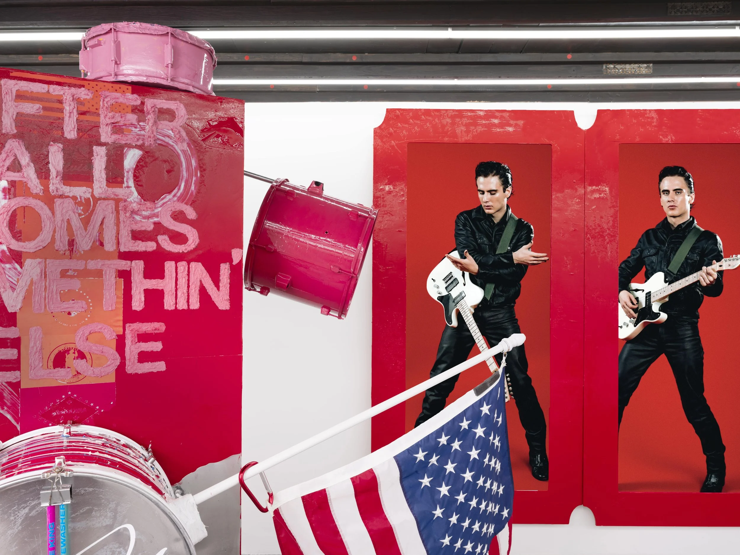 Two men dressed in black leather jackets and pants, holding electric guitars, standing in front of red wall panels with their guitars at their sides. There is a blue and white American flag on a pole in front of them. To the left, there is a pink drum, a red metal trash can, and pink painted text on a pink wall that reads "Better All Homes Within".