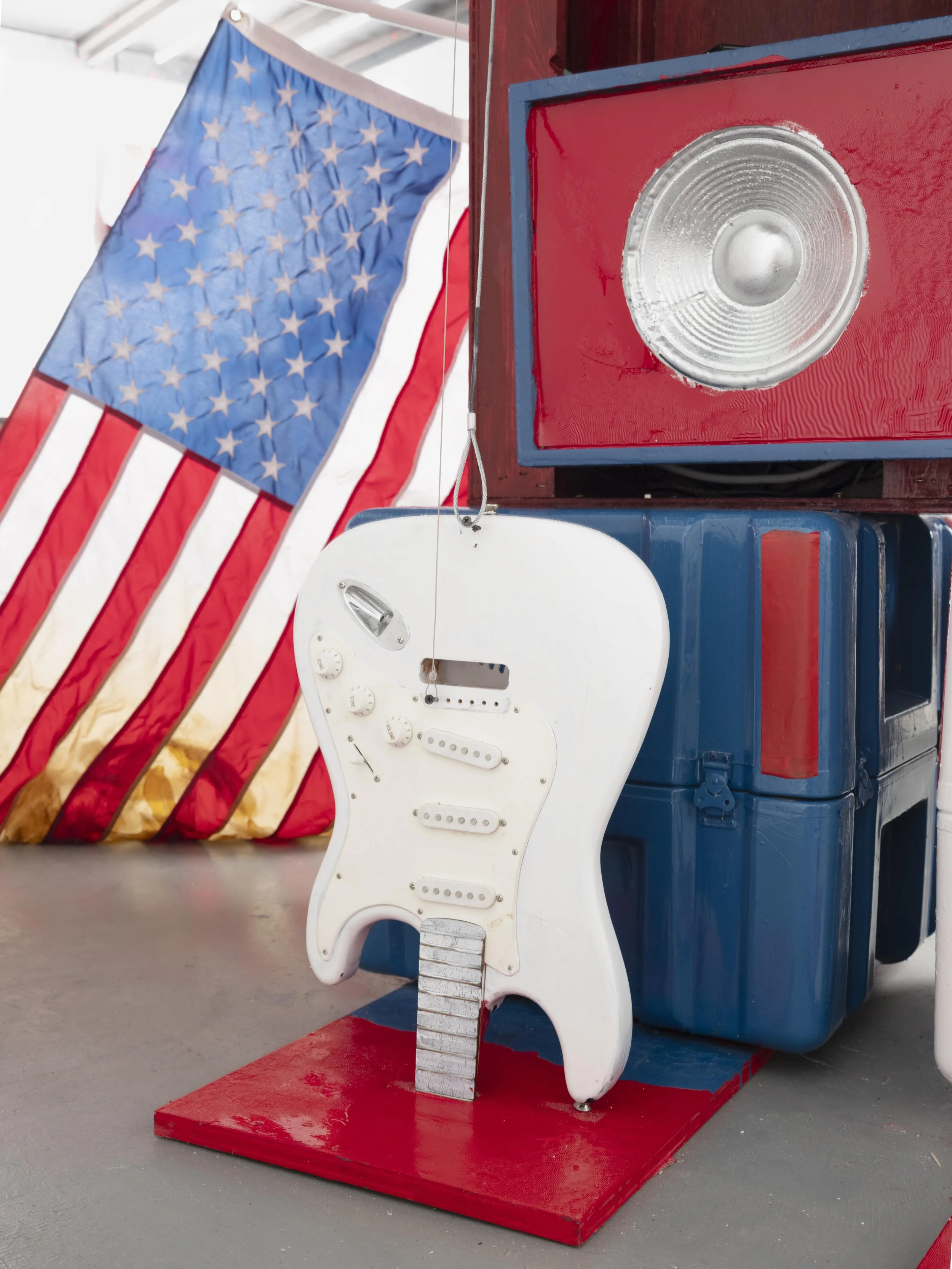 A white electric guitar replica positioned on a red platform, in front of a large blue plastic container, with a vintage speaker mounted on a red and blue box. An American flag in the background.