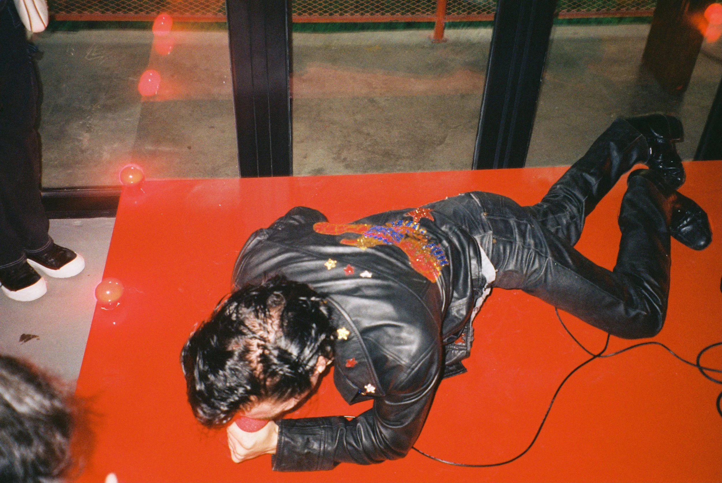Person lying on their side on a red surface, wearing a black leather jacket with embroidery, black pants, and shoes, with hair covering their face, in front of glass doors with reflections of red lights and a person in the background.