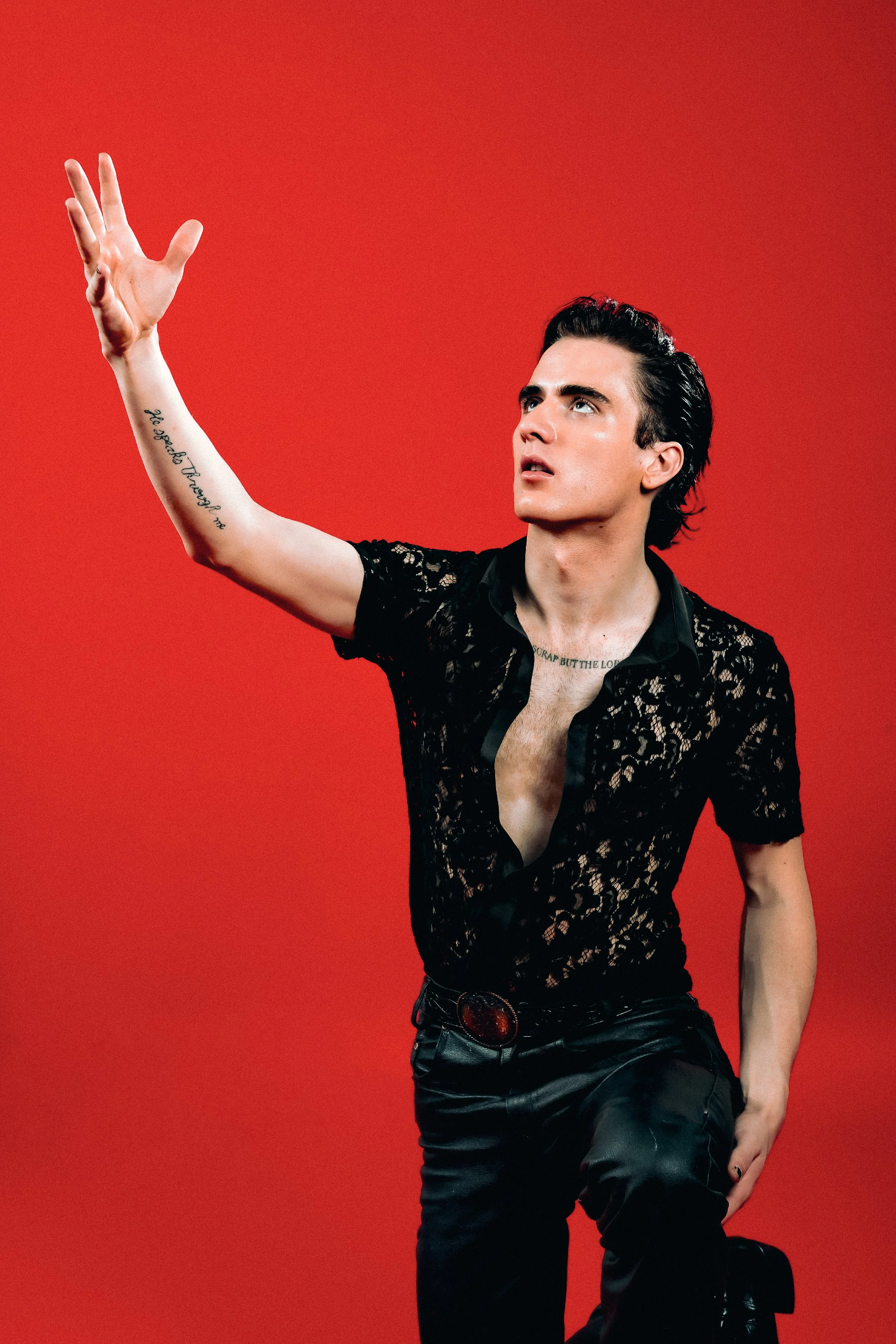 A young man with dark hair styled back, wearing a black lace shirt partially unbuttoned, black leather pants, and a belt with a large buckle, is kneeling on one knee against a red background. He is raising his right arm with an open hand and looking upwards.