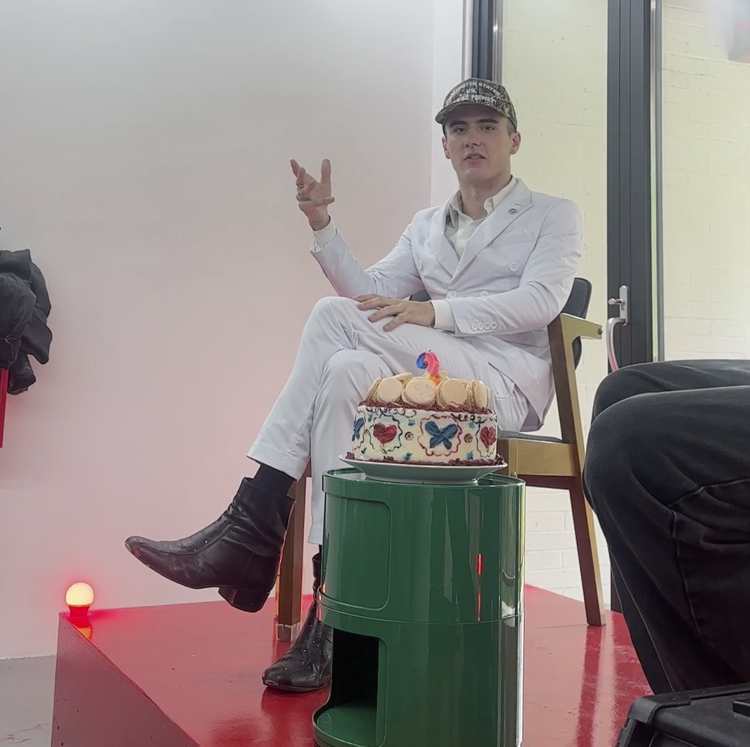 Man in white suit and camouflage cap sitting on a chair with one leg crossed over the other, gesturing with his hand, in front of a decorated birthday cake with macarons and a candle. The cake is on a green stand, placed on a red platform inside a room with glass doors and white walls.