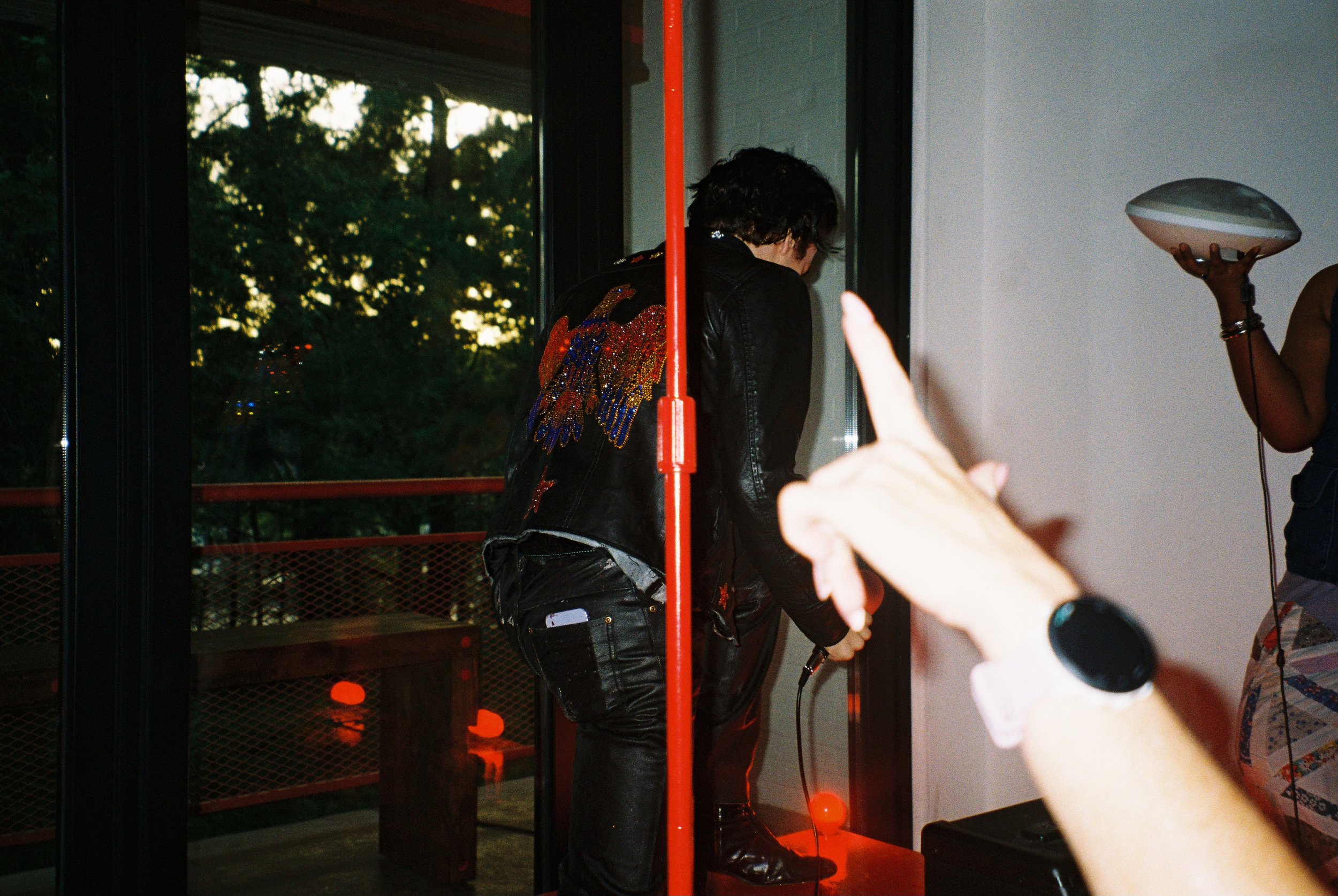 A person with dark, curly hair wearing a black leather jacket with colorful embroidered designs on the back, standing near a glass door with a red pole in the foreground, inside a room with a white brick wall, a person holding a white device, and a hand pointing towards the person with the embroidered jacket.