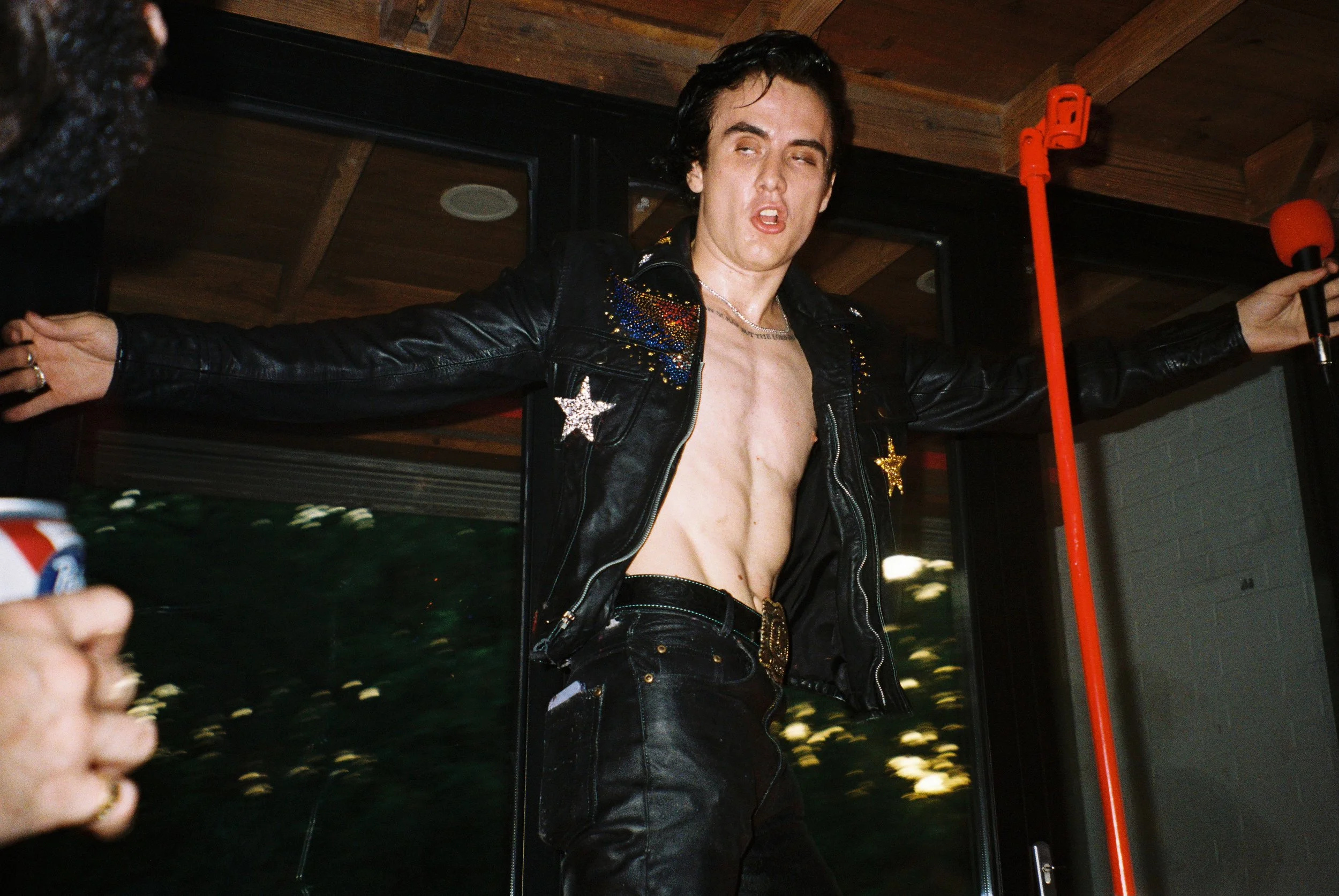 A man with curly dark hair and light skin is wearing a black leather jacket with colorful embellishments and a star patch, unzipped to reveal his bare chest, and black leather pants, standing with arms outstretched inside a room with a wooden ceiling and large windows. He appears to be performing or posing.