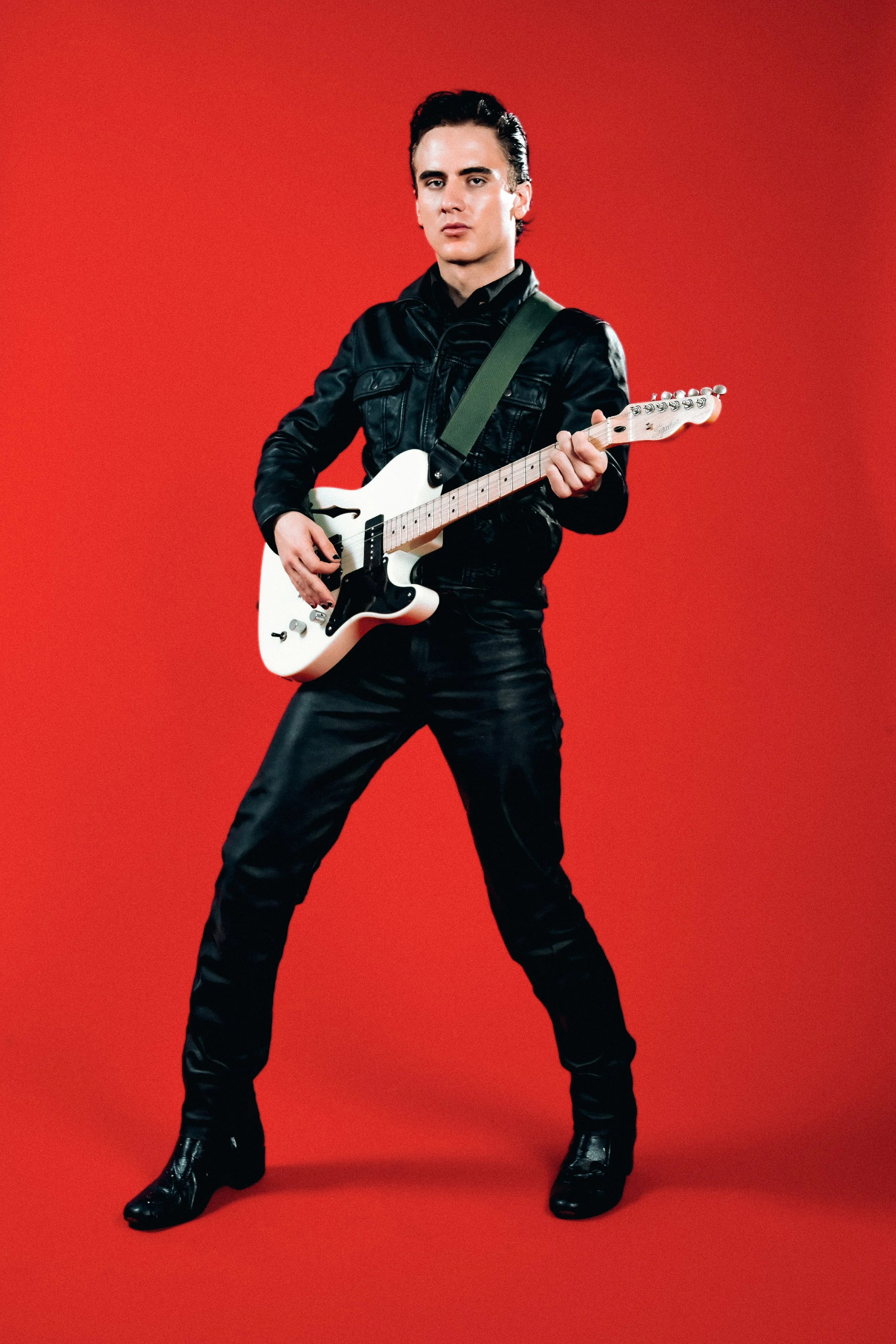 A young man in black leather clothing playing a white electric guitar against a red background.