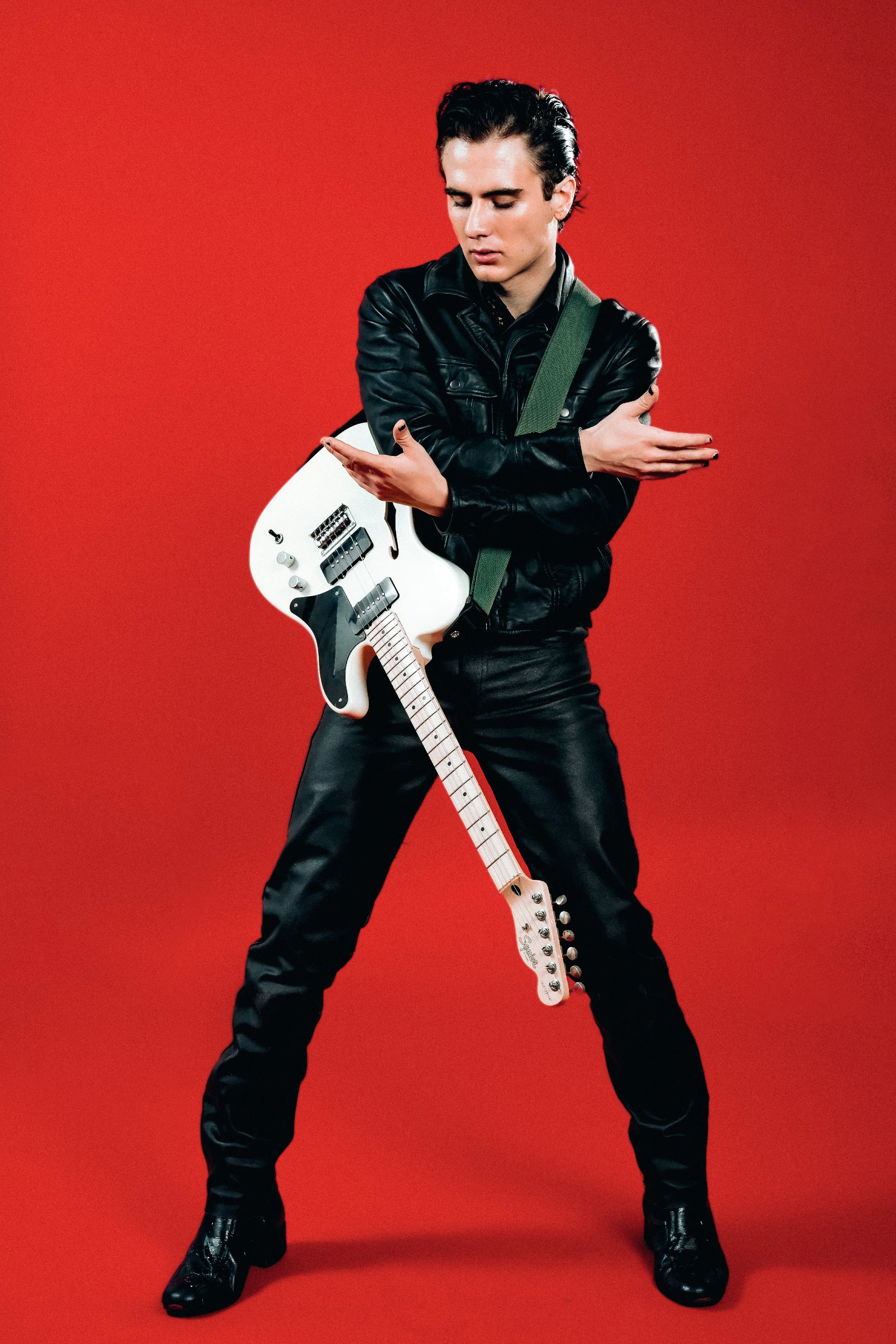 A young man stands against a red background, holding an electric guitar while crossing his arms, with a serious expression.