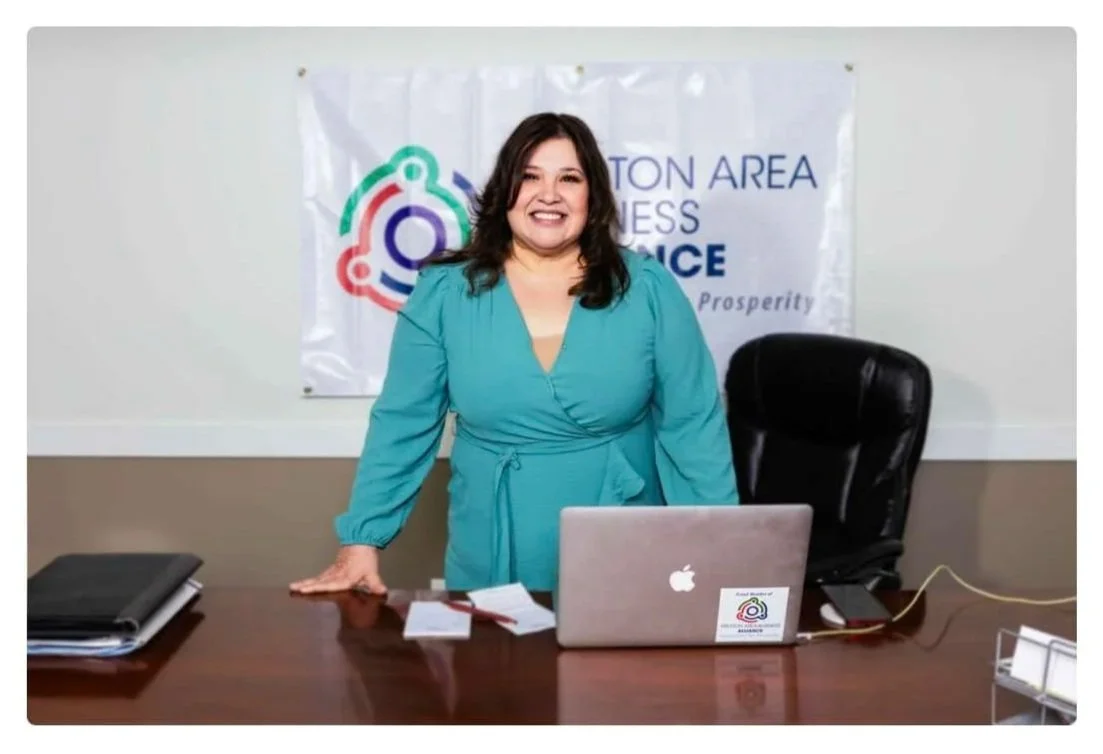 Woman in turquoise dress standing behind a desk with a laptop, in an office setting with a banner that reads 'HOUSTON AREA BUSINESS CONFERENCE' behind her.