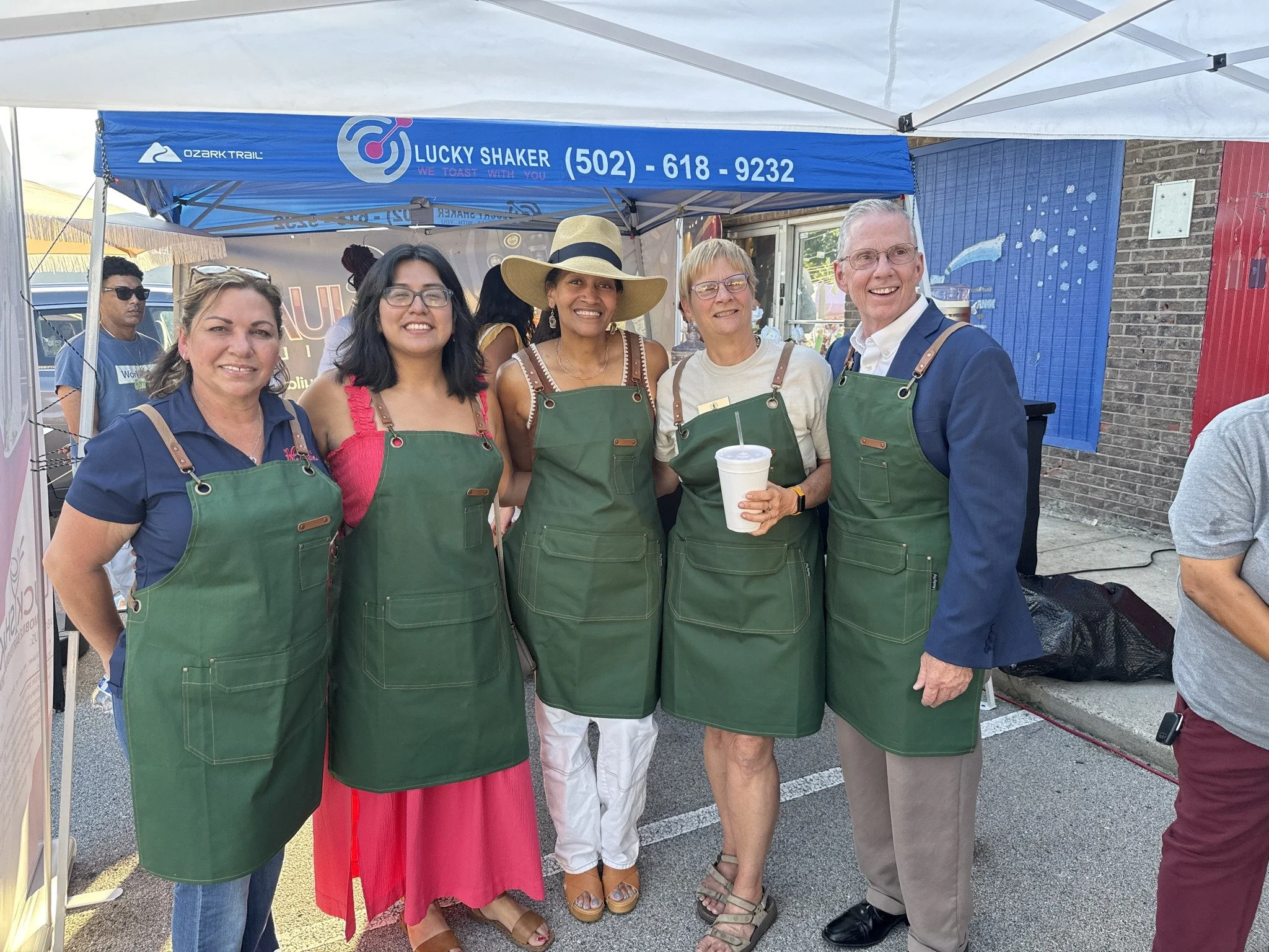 Five people wearing green aprons standing together outdoors at a festival or fair, smiling at the camera, with one holding a drink.
