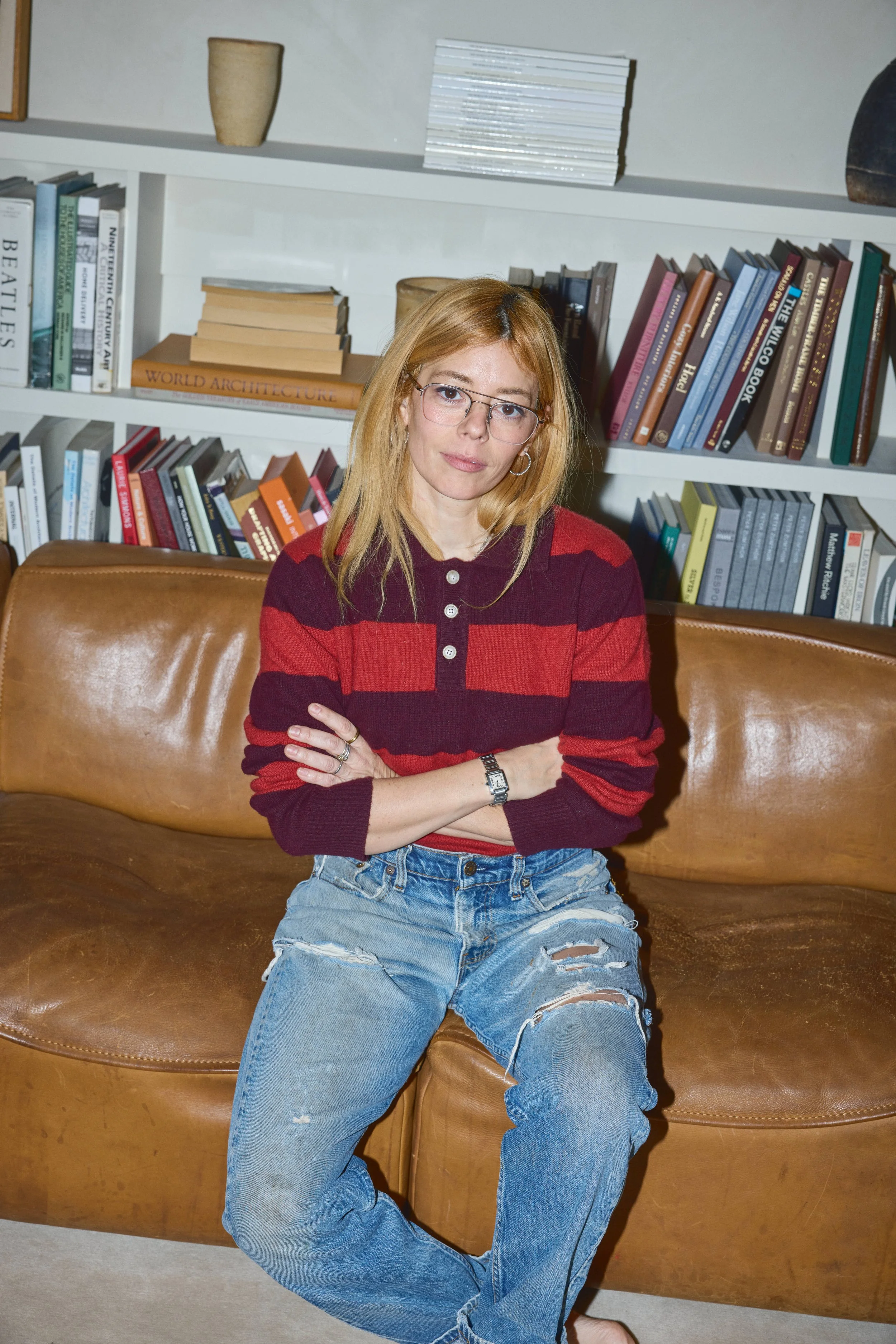 A woman with glasses and blonde hair sitting on a brown leather couch in front of a bookshelf filled with books in a cozy room.