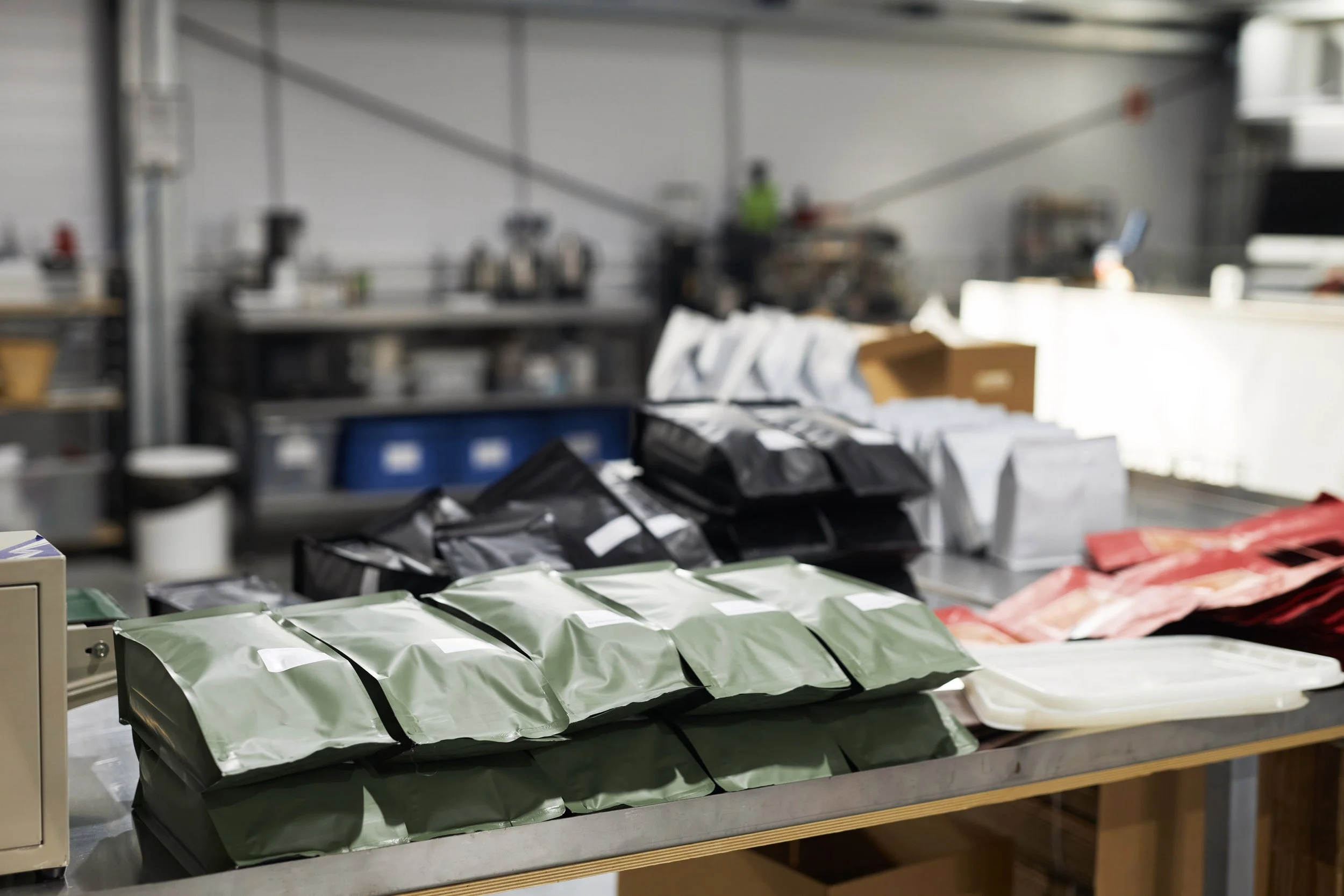 A metal table with multiple vacuum-sealed packages in various colors, including green, black, white, and red, arranged in rows inside a workshop or food production facility.