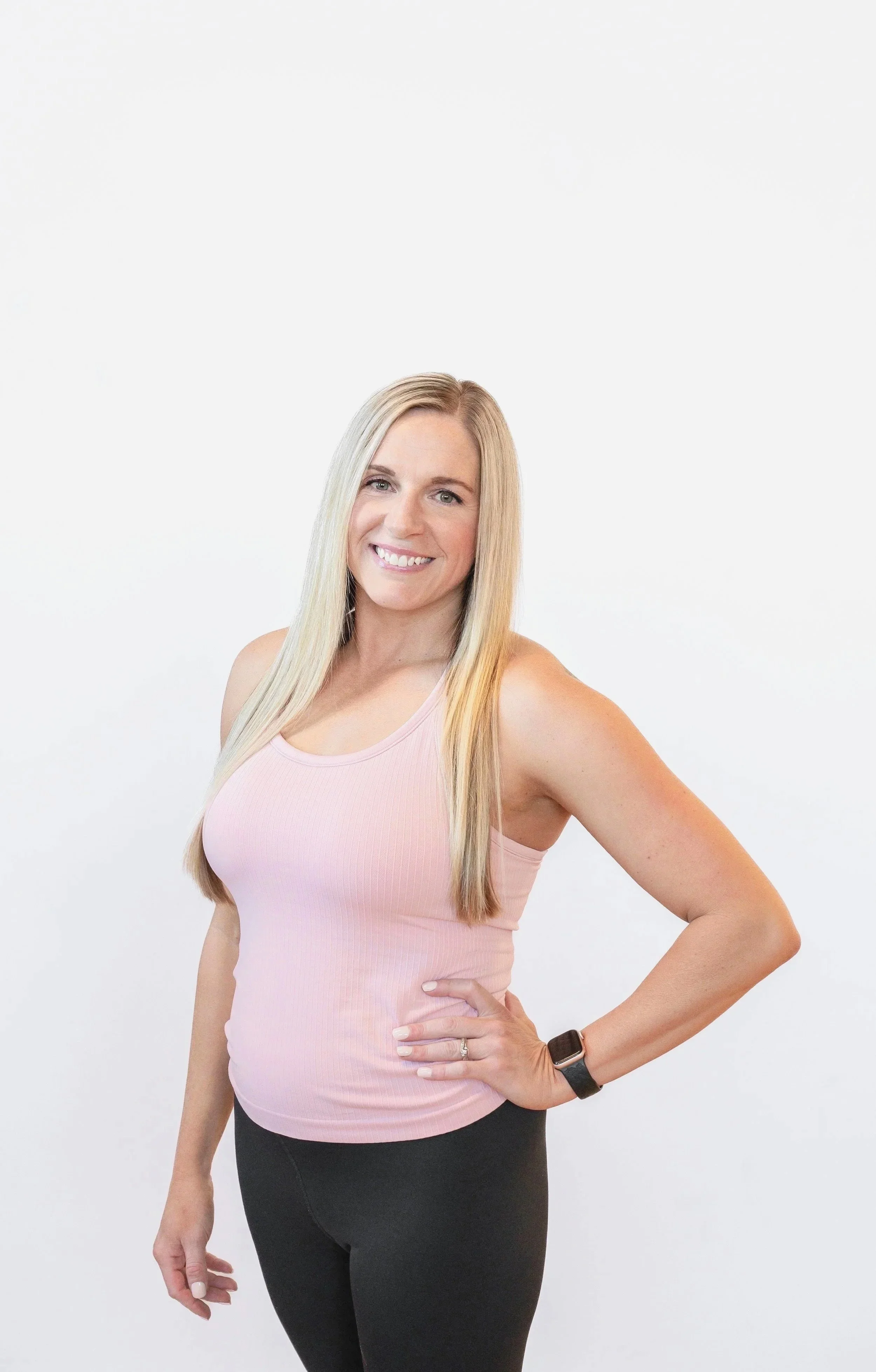 A woman smiling, with long blonde hair, wearing a pink tank top and a smart watch on her left wrist, standing against a plain light background.
