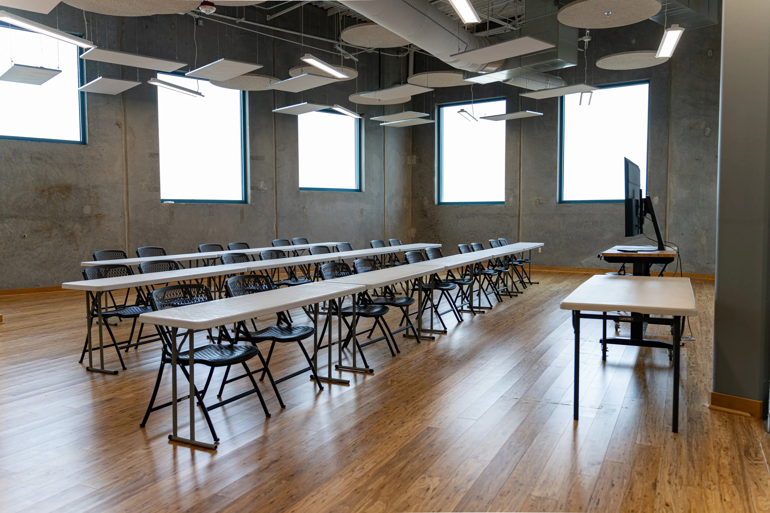 Empty modern classroom or conference room with multiple white tables and black chairs arranged in rows. Large windows on the back wall let in natural light. A computer monitor is placed on a small desk at the front of the room.