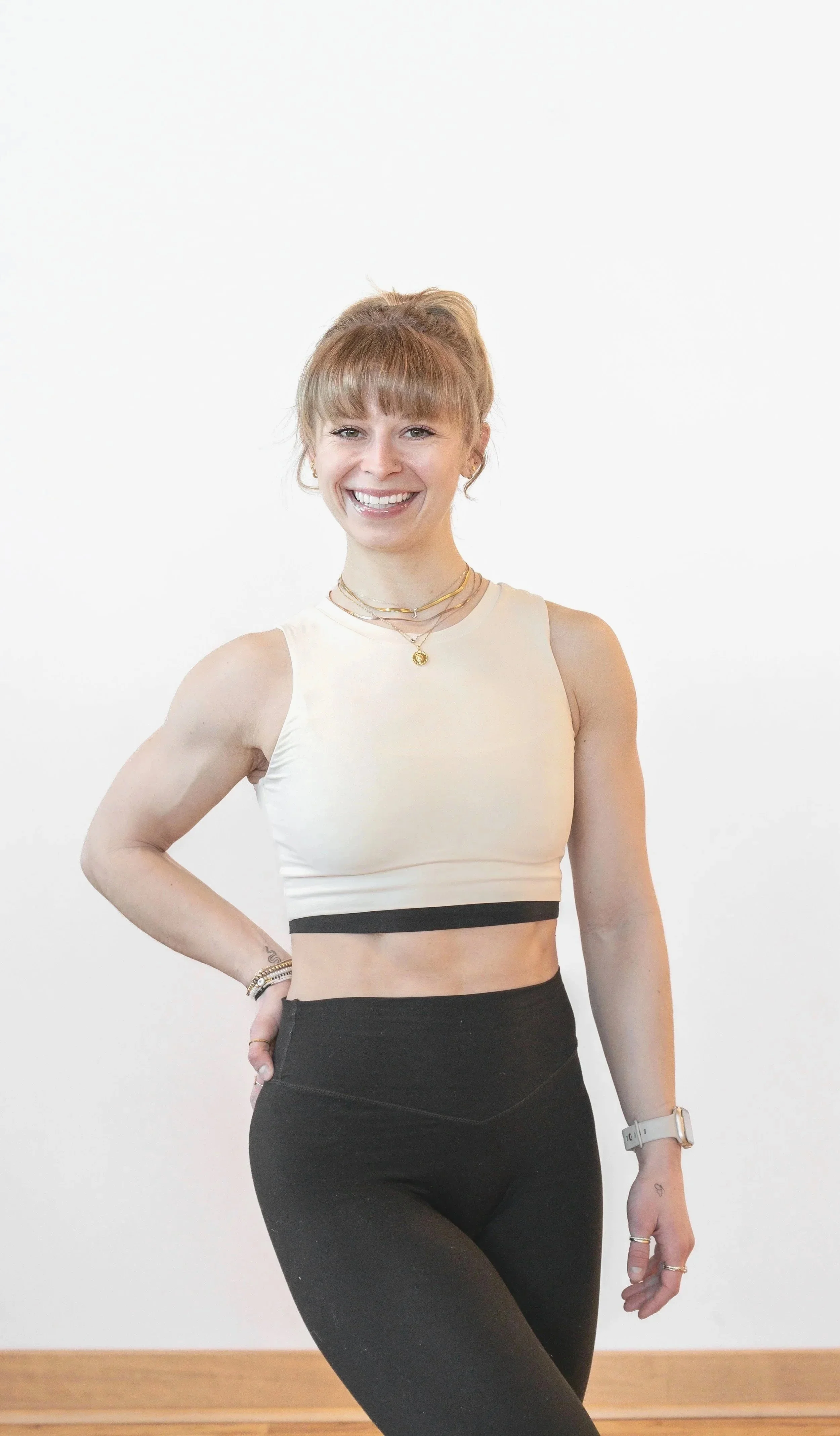 A young woman with blonde hair and a bright smile, wearing a white crop top and black athletic pants posing with her hand on her hip against a plain light-colored background.