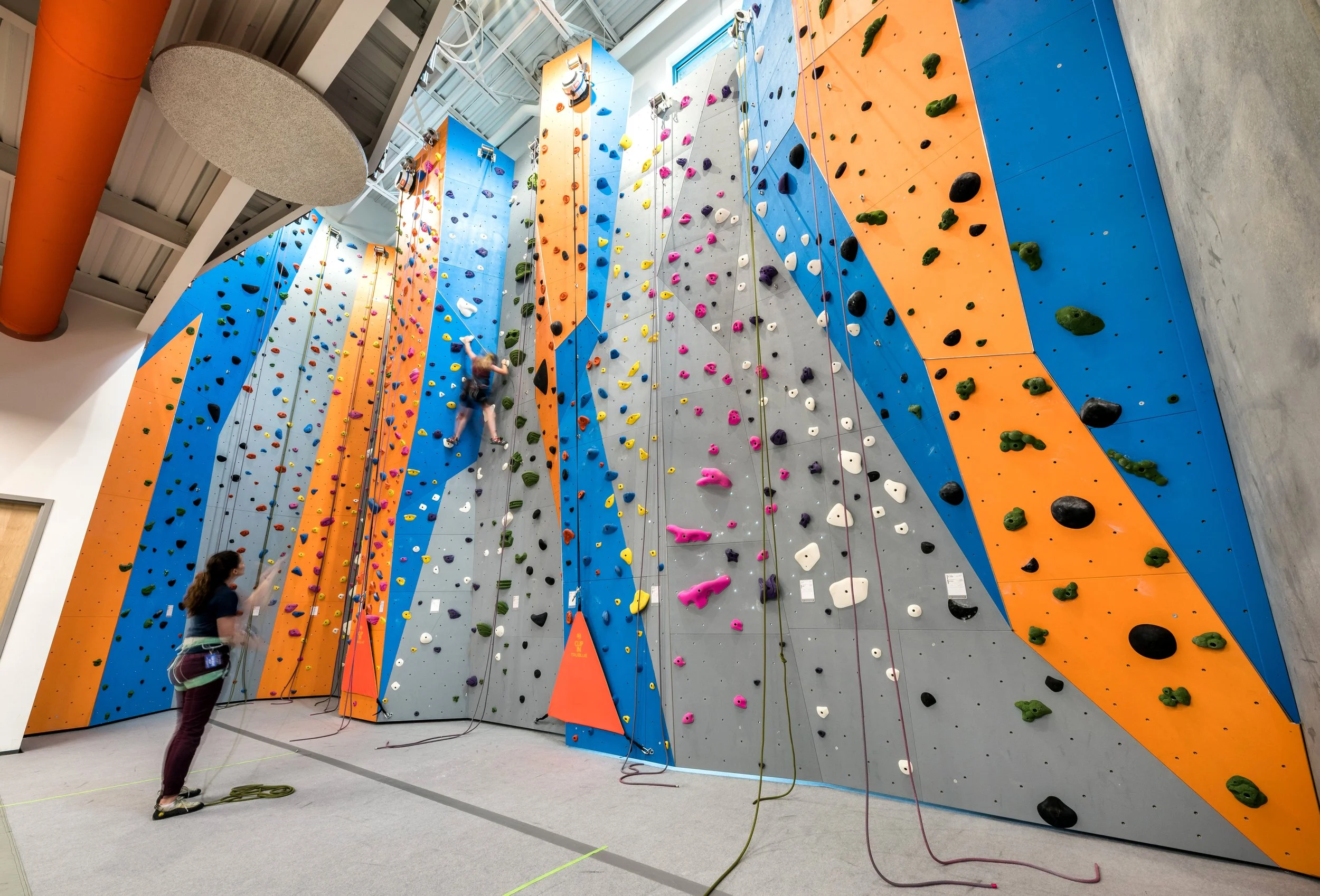 Indoor rock climbing gym with colorful climbing walls and a person climbing while an instructor spotting. The walls are decorated with various holds in different colors and shapes.