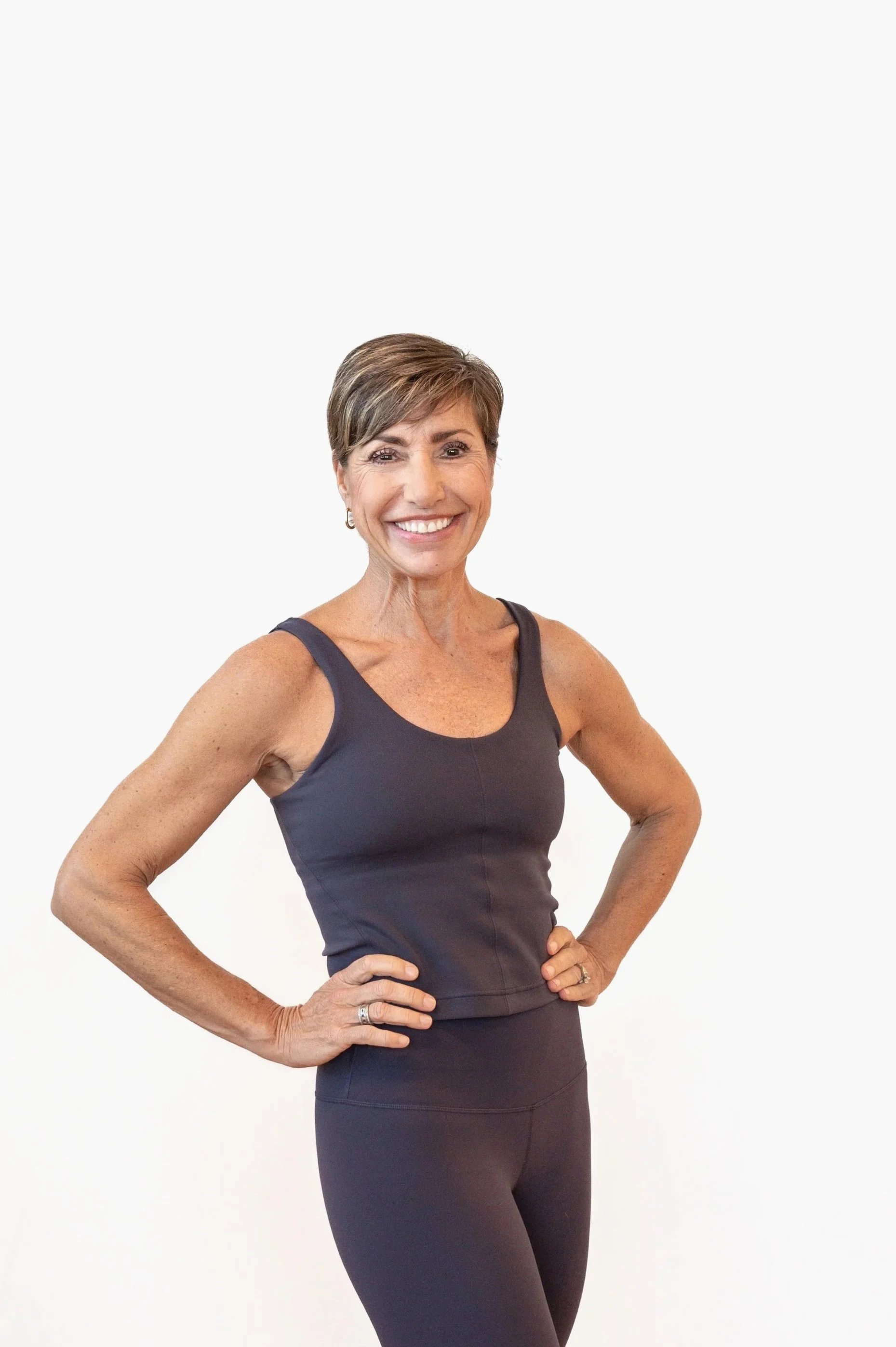 A smiling middle-aged woman standing against a plain light-colored wall, wearing a purple athletic tank top and matching leggings.