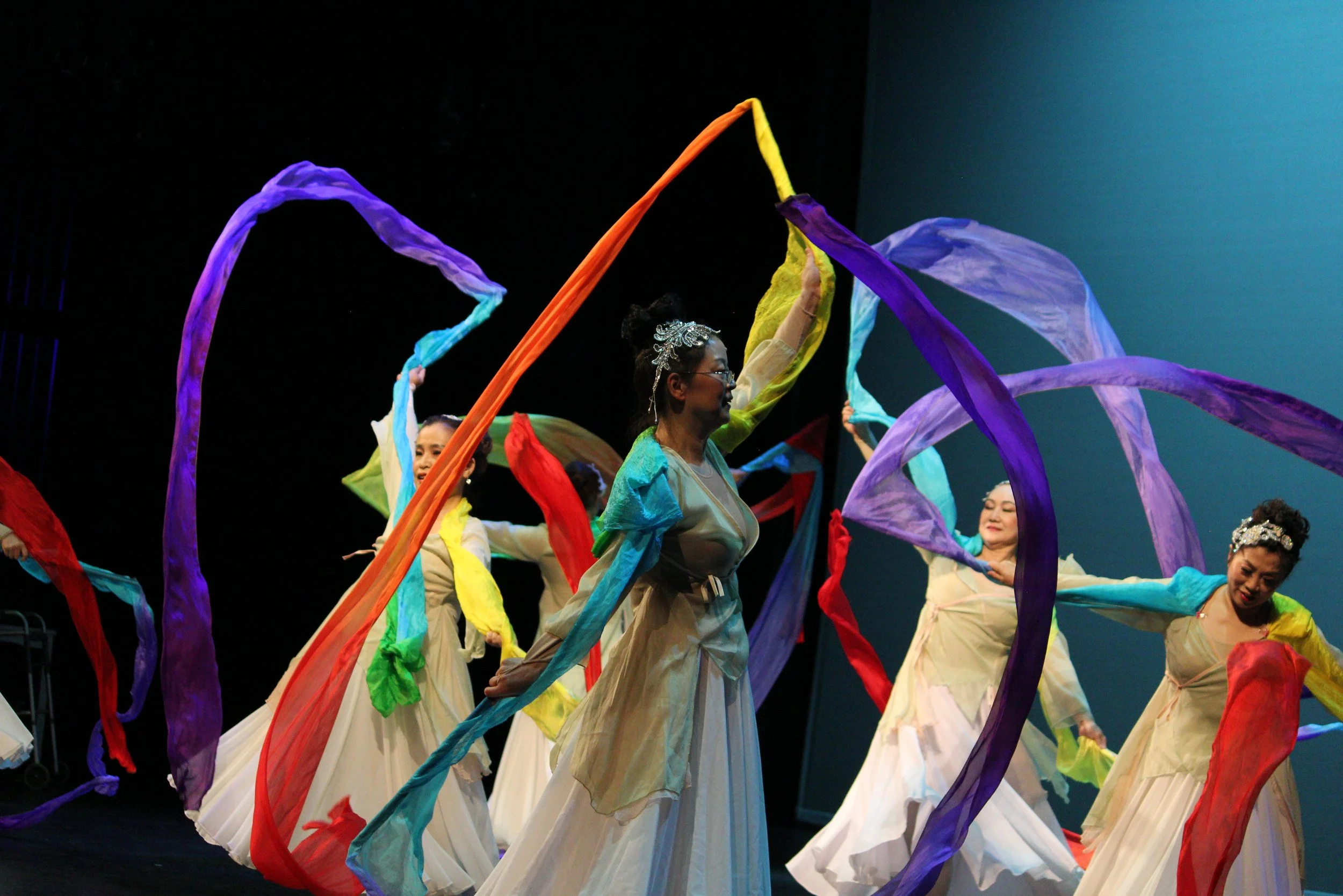 A group of older dancers from the Melody Institute perform a rainbow ribbon dance on stage. 