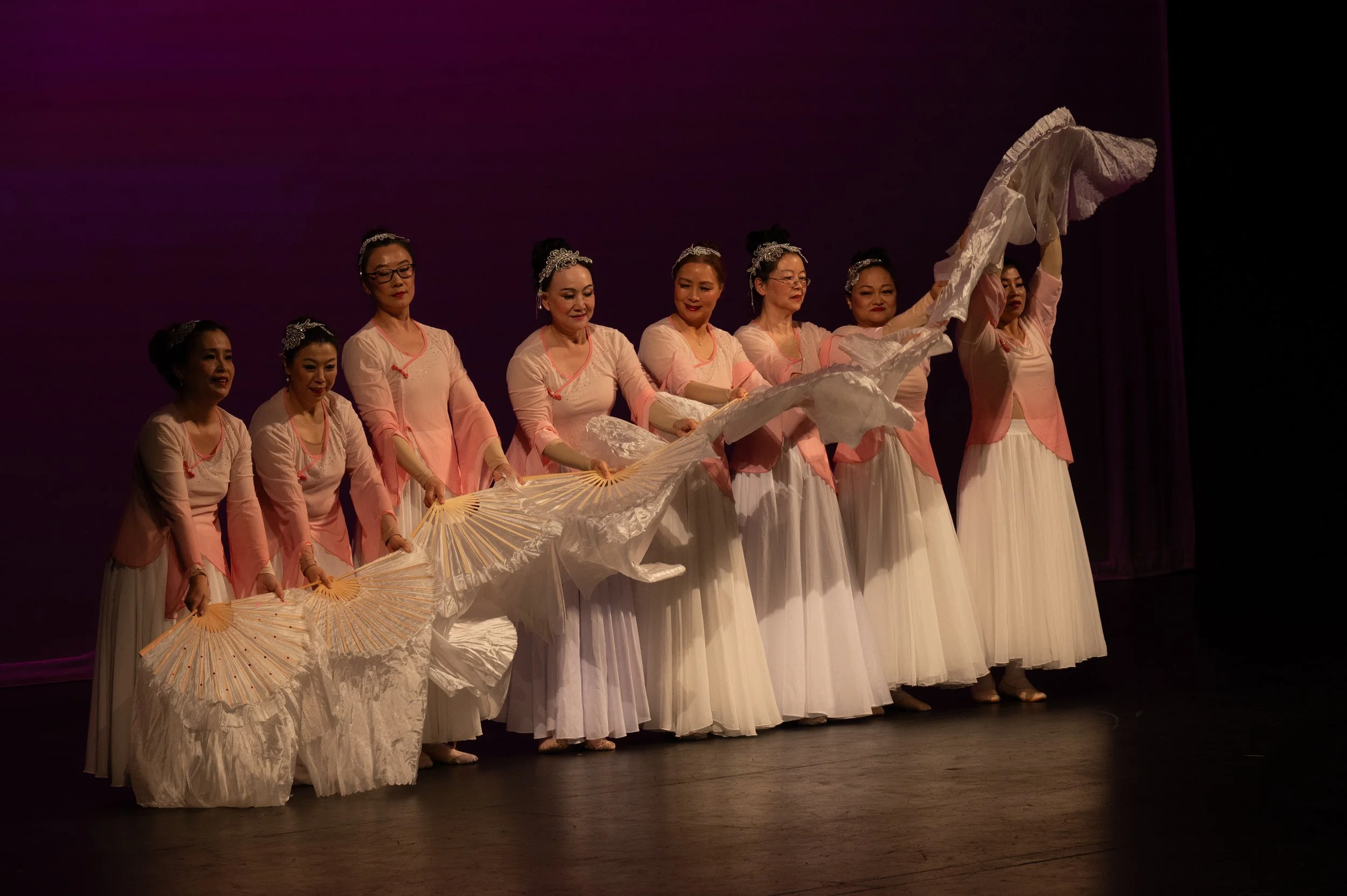 A group of older dancers from the Melody Dance Institute performing a fan wave on the stage