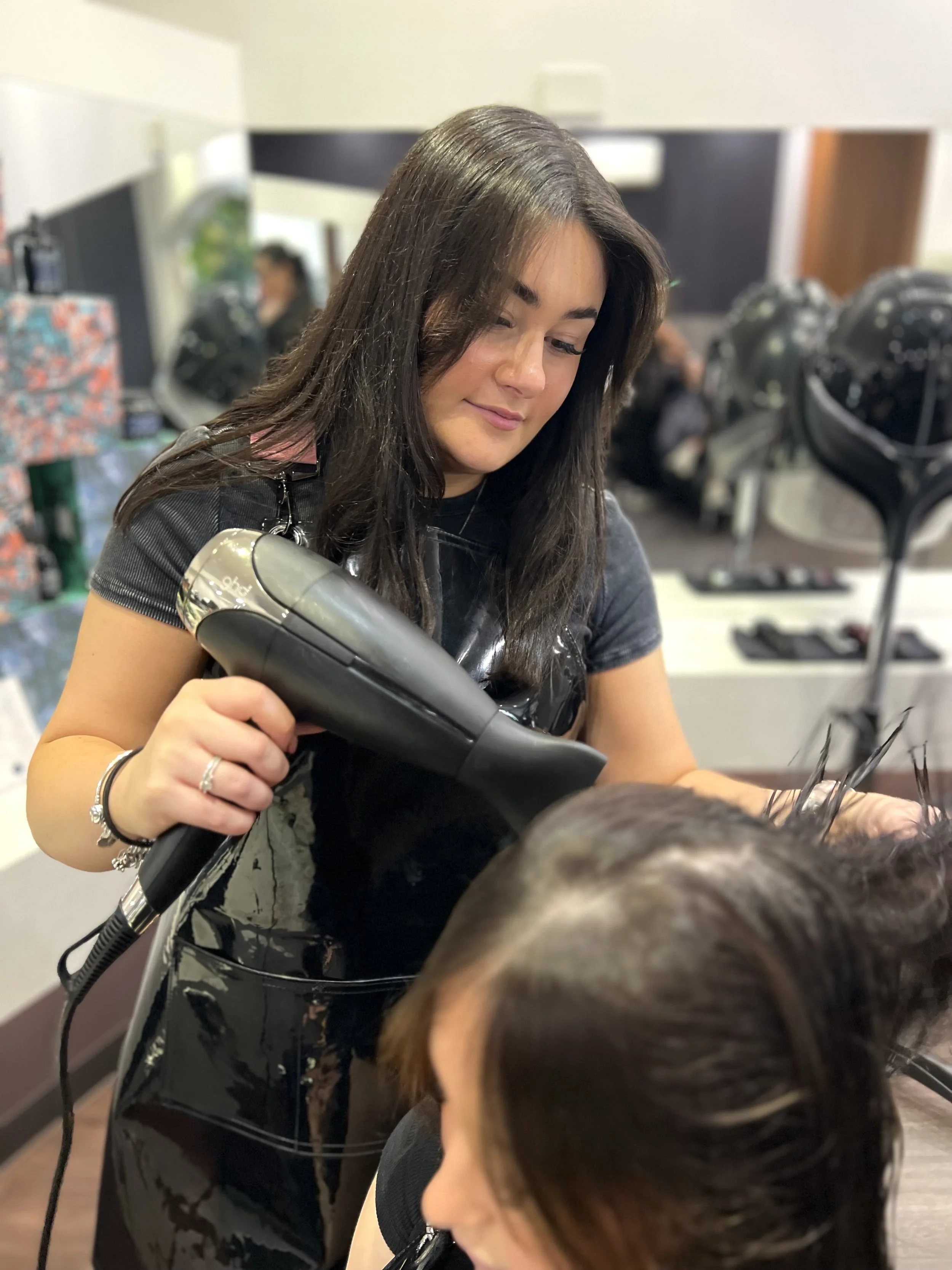 A hairstylist dries a woman's hair with a black hairdryer in a salon.