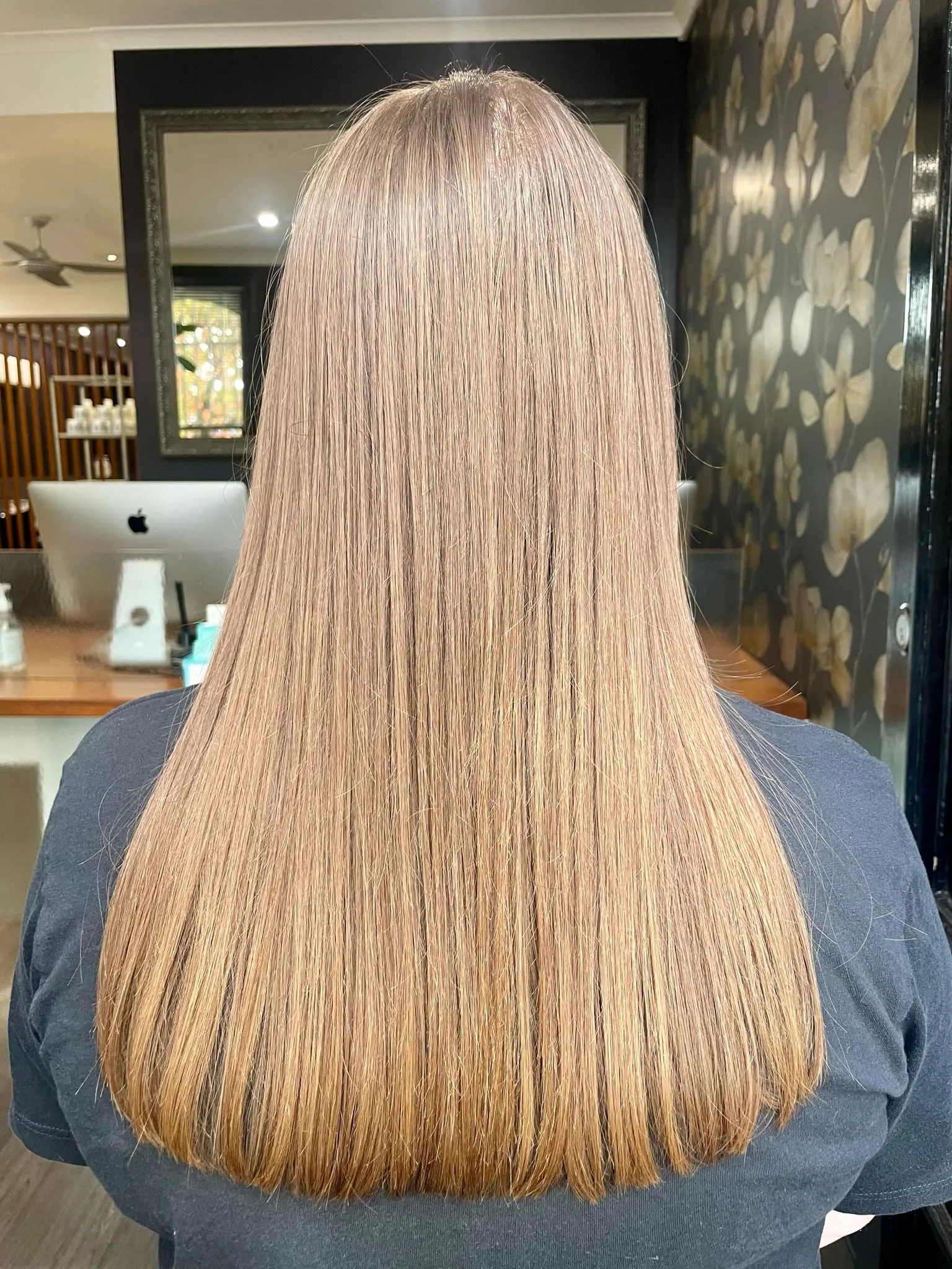 Back of a woman with straight, shiny, light blonde hair, cut to shoulder length, sitting in a modern salon with dark wall and floral wallpaper.