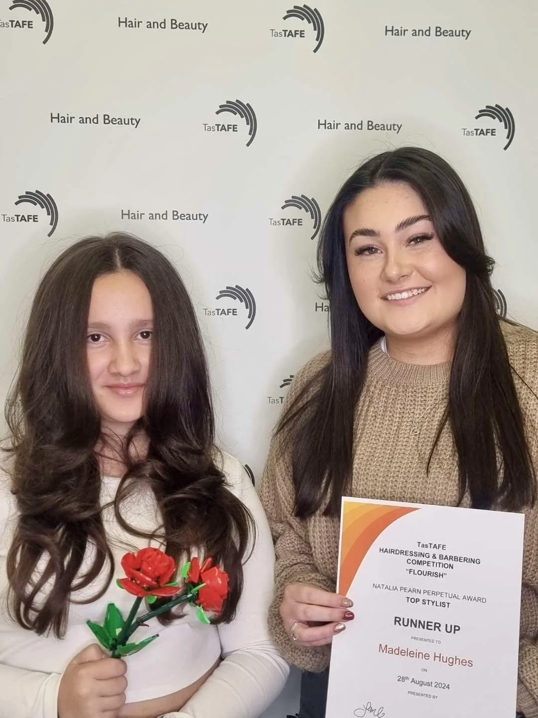 Two women posing for a photo at a hair and beauty competition event. The woman on the left has long, dark curly hair and is holding a small red and green plastic flower. The woman on the right, with long dark straight hair, is holding a certificate f