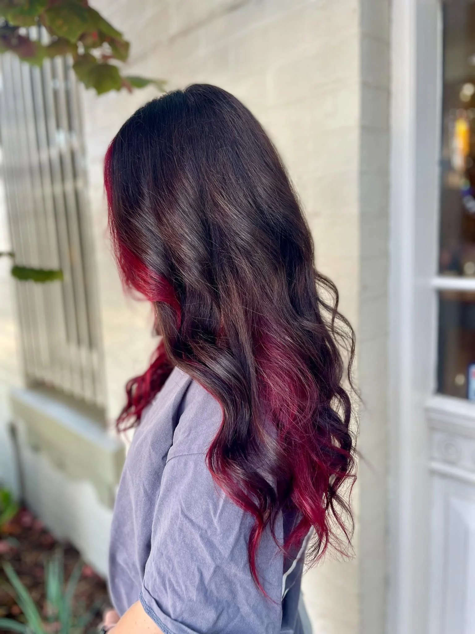 A woman with long, wavy black hair with pink highlights standing outdoors next to a beige wall and a window.