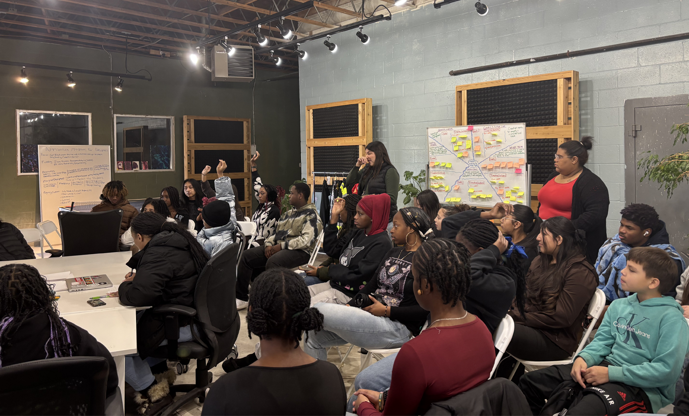 Group of young people attending a workshop or presentation in a room with gray and green walls, some raising their hands, with two women standing near a whiteboard covered with colorful sticky notes.