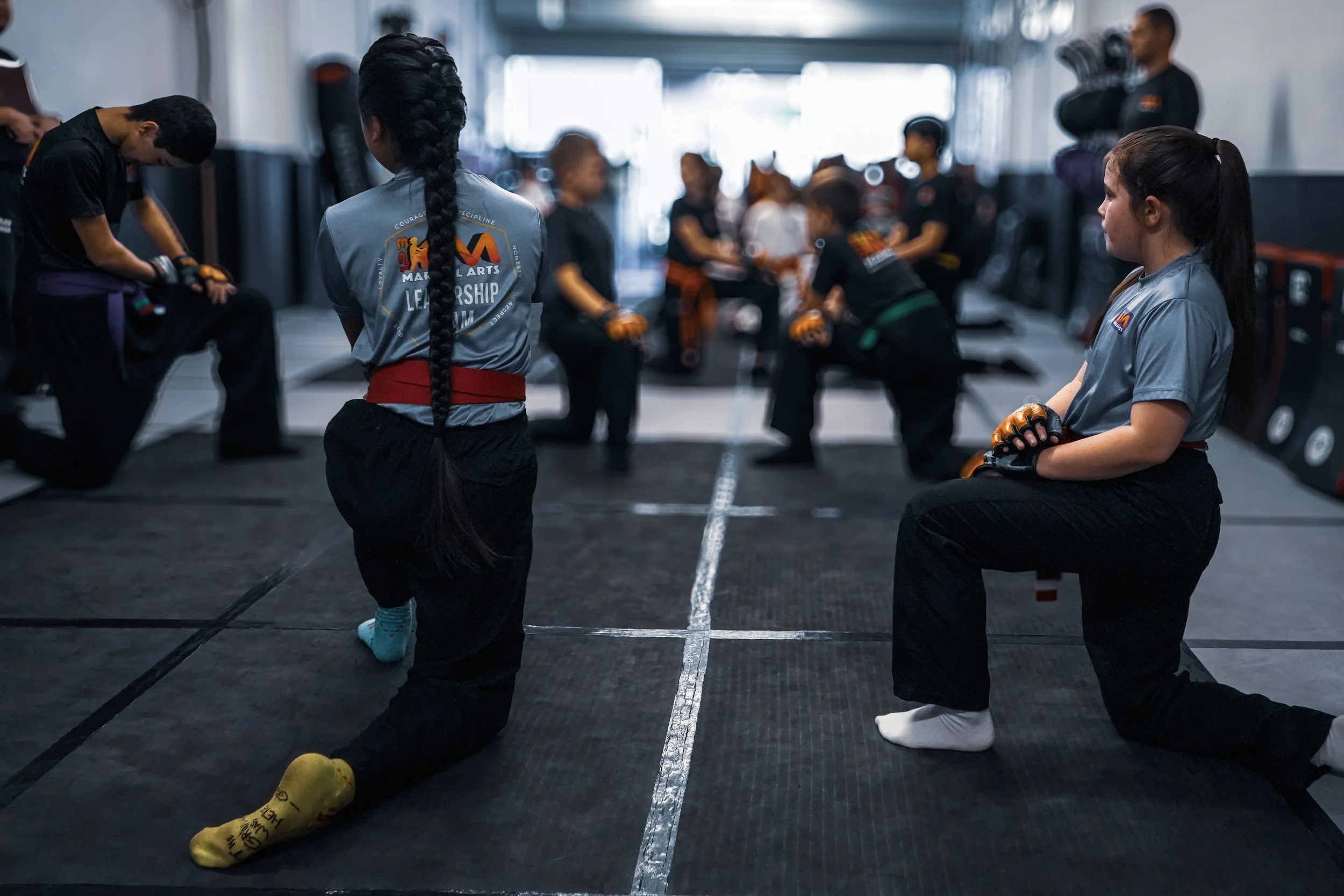 Martial arts students in a gym practicing and listening to instructor, some in traditional gi with belts, some sitting on the floor with gloves, and a coach in the background holding pads.
