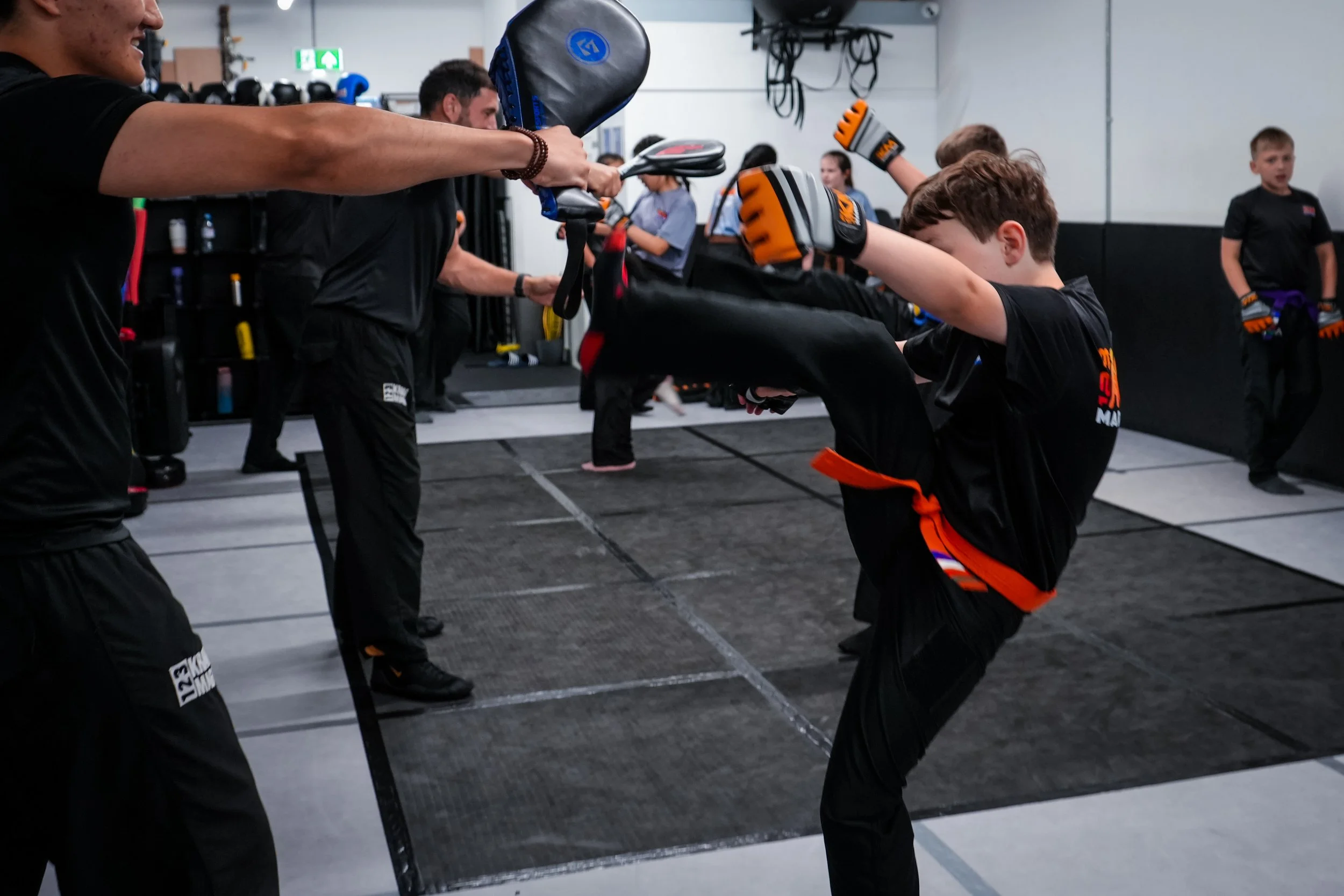 Kids practicing martial arts or kickboxing exercises in a gym, wearing gloves and black uniforms, with adults instructing them.