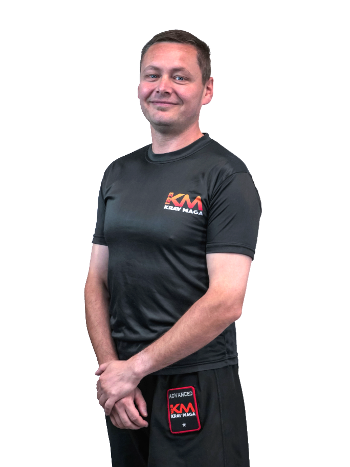 A young man with short brown hair smiling at the camera, wearing a black t-shirt and black shorts, both branded with Krav Maga logos, standing with hands clasped in front of him against a plain white background.