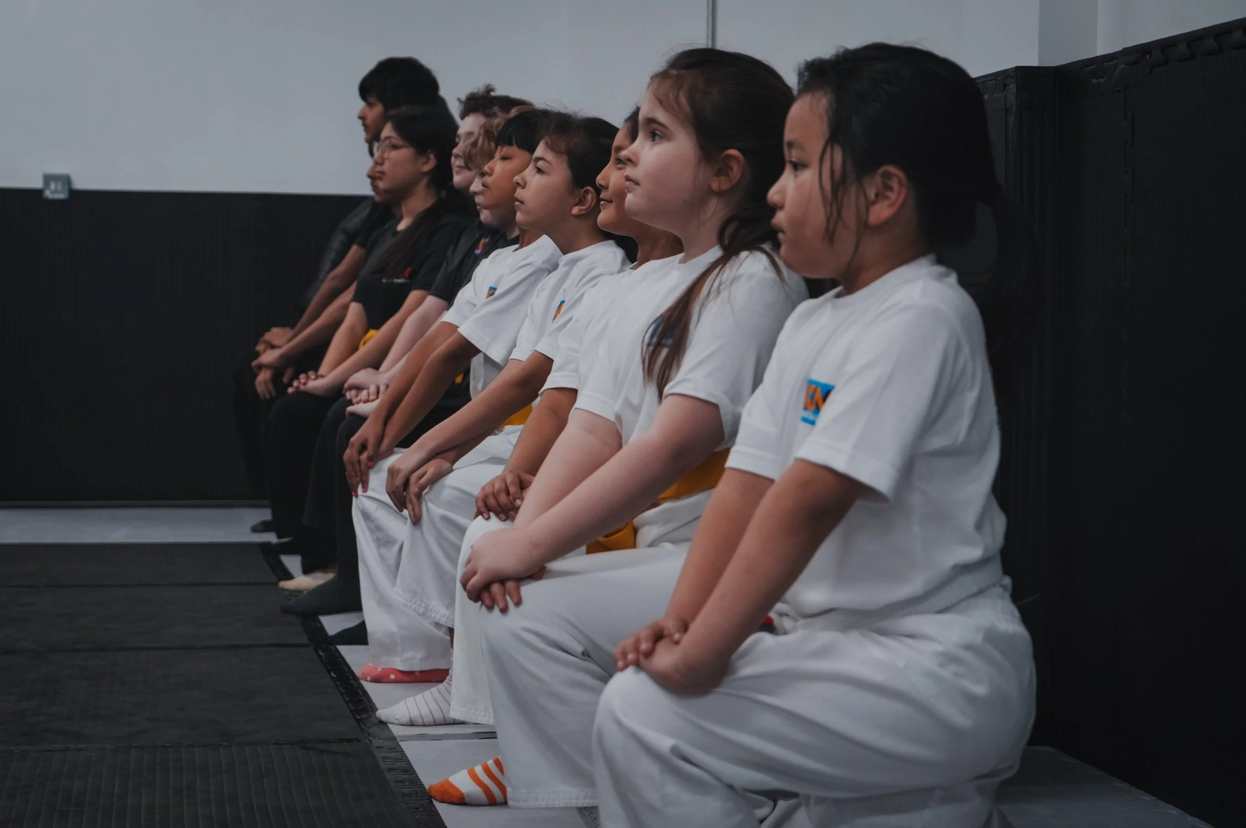 Children sitting cross-legged in a row, dressed in martial arts uniforms, inside a gym or dojo, paying attention.