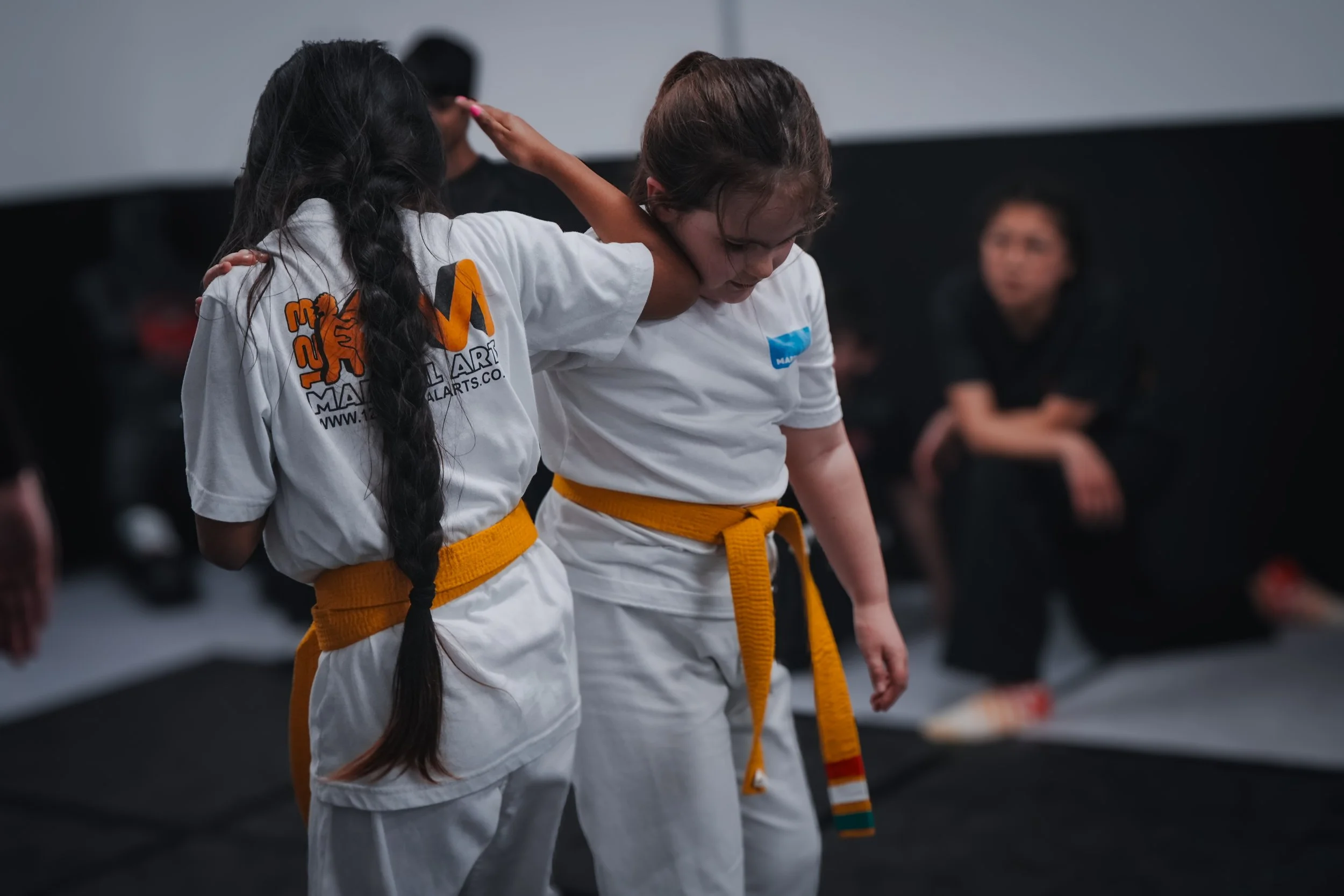 Two martial arts students, wearing white gis with yellow belts, interact in a training room, with other students and a monitor in the background.