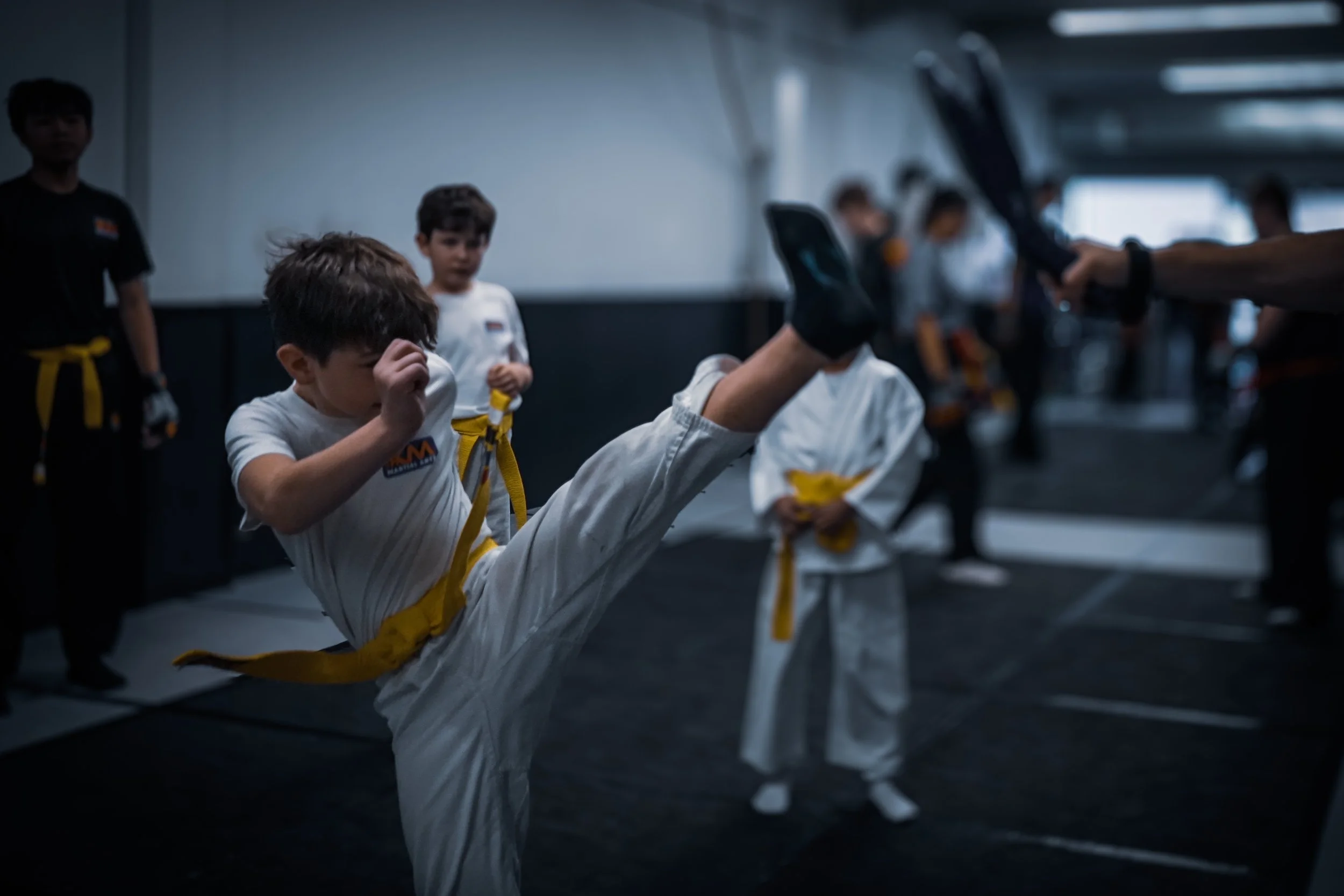 Children in martial arts uniforms practicing a high kick in a training room.