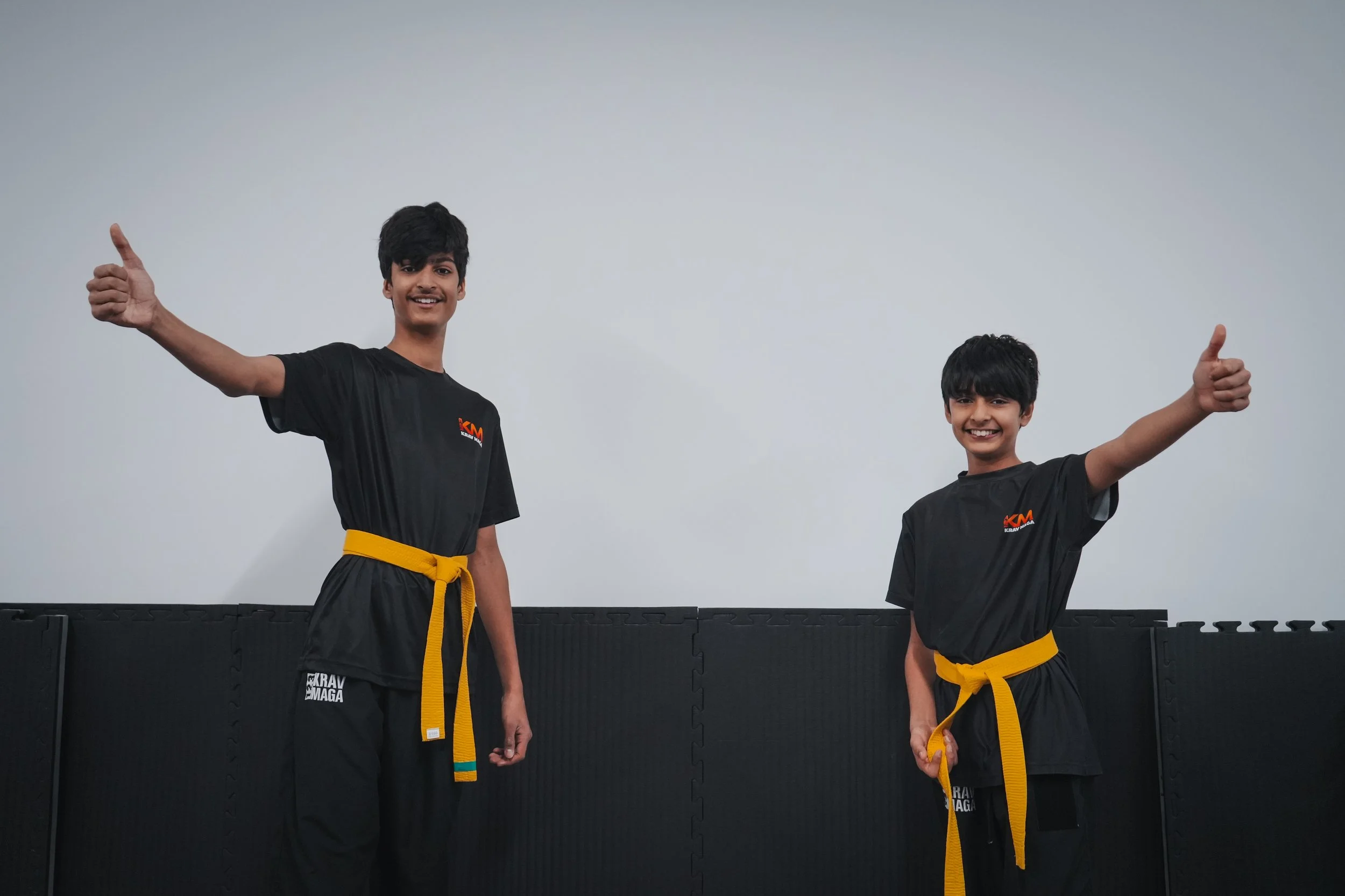 Two children dressed in judo uniforms with different colored belts, smiling and giving thumbs up.
