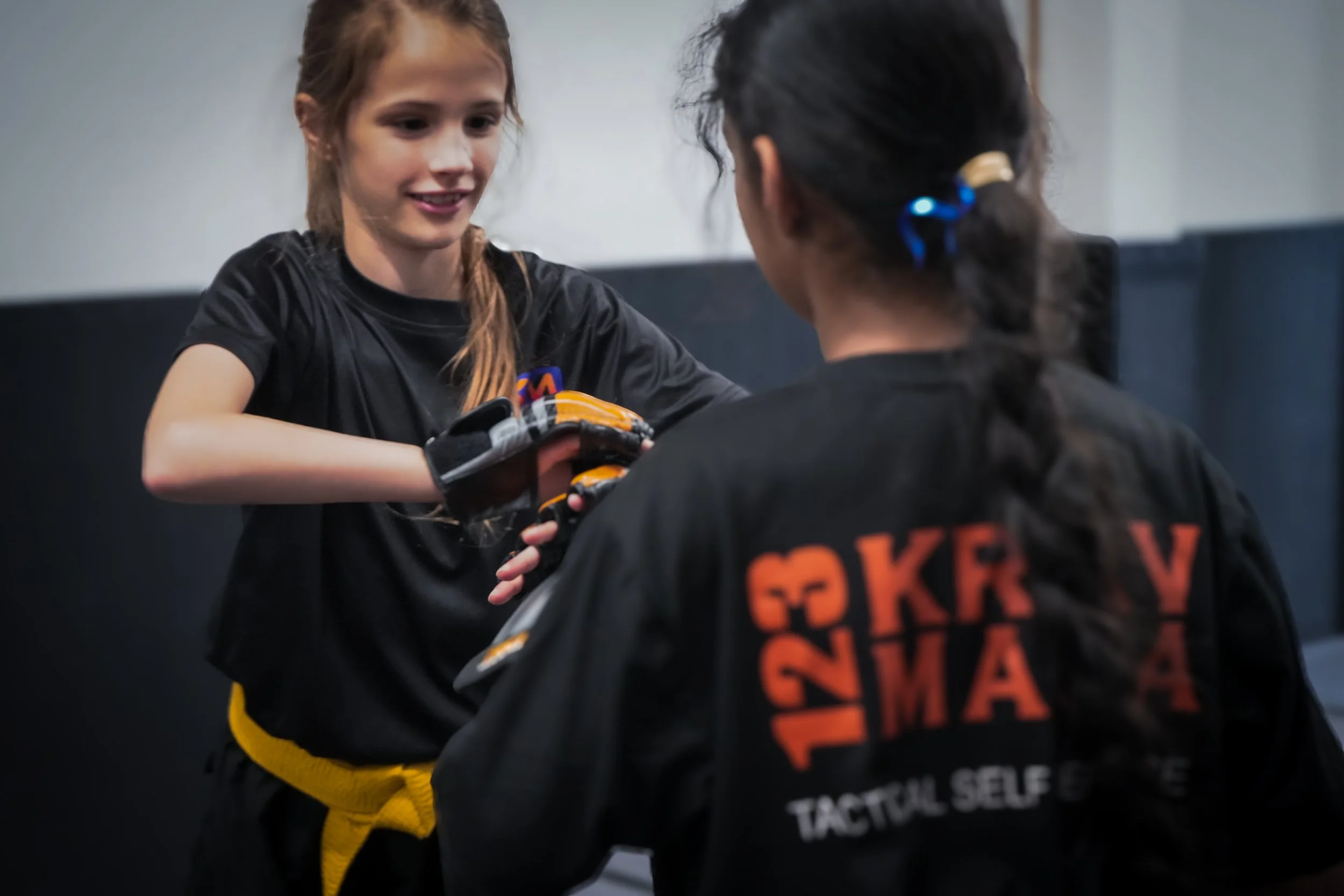 A young girl with red hair in a braid, wearing a black t-shirt and yellow martial arts uniform, smiling as she helps an instructor with black hair tied back, wearing a black gi and an orange belt, during martial arts training. The girl is holding a punching pad while the instructor is adjusting her gloves.
