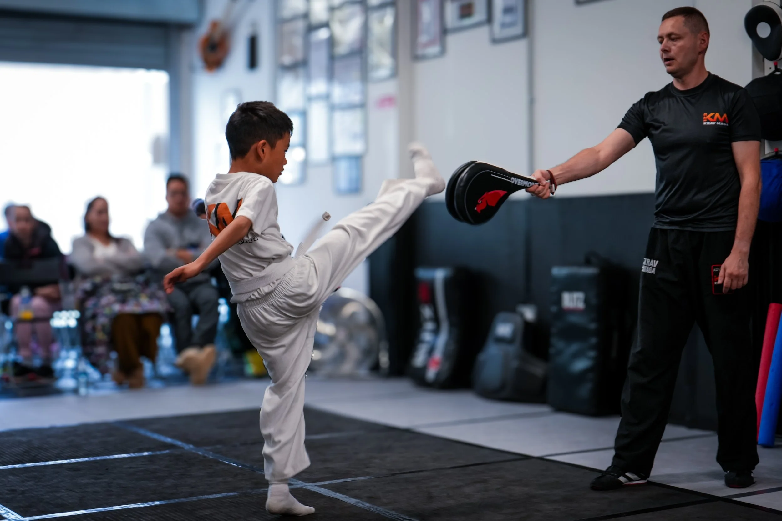A young boy in a martial arts uniform practicing a high kick while a male instructor in sportswear holds a pad for him in a martial arts gym. Several people are seated in the background.