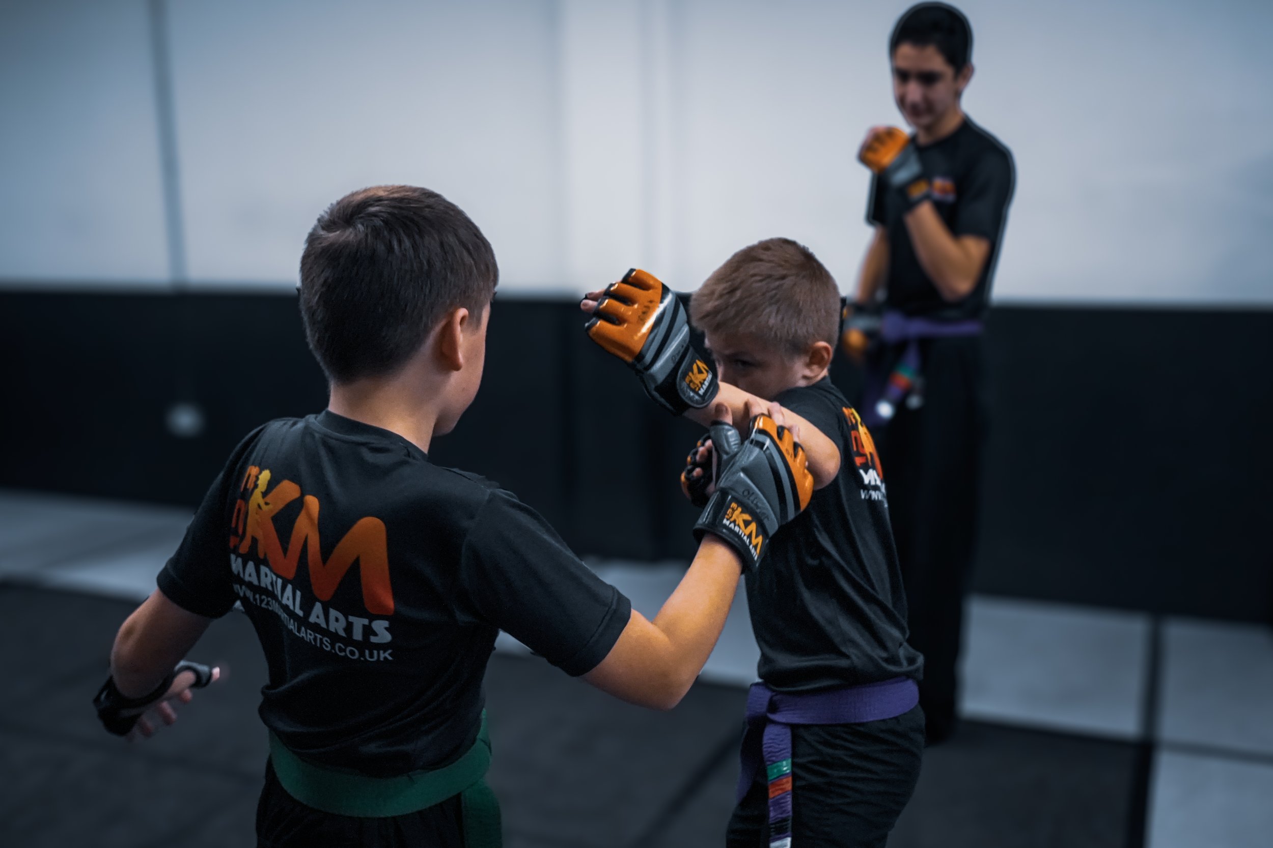 Two young martial artists sparring, wearing black uniforms with orange and purple belts, in a gym with a coach observing in the background.