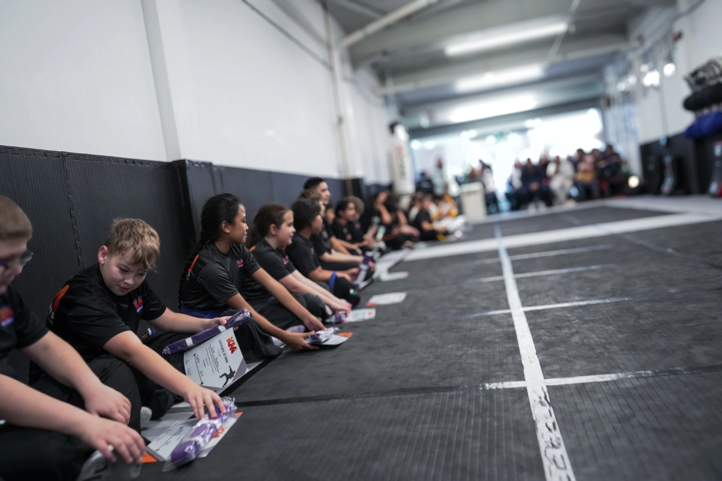 A group of children sitting on the floor in a gym, wearing black uniforms, each holding a certificate and a small gift bag, with a row of spectators and vehicles visible in the background.