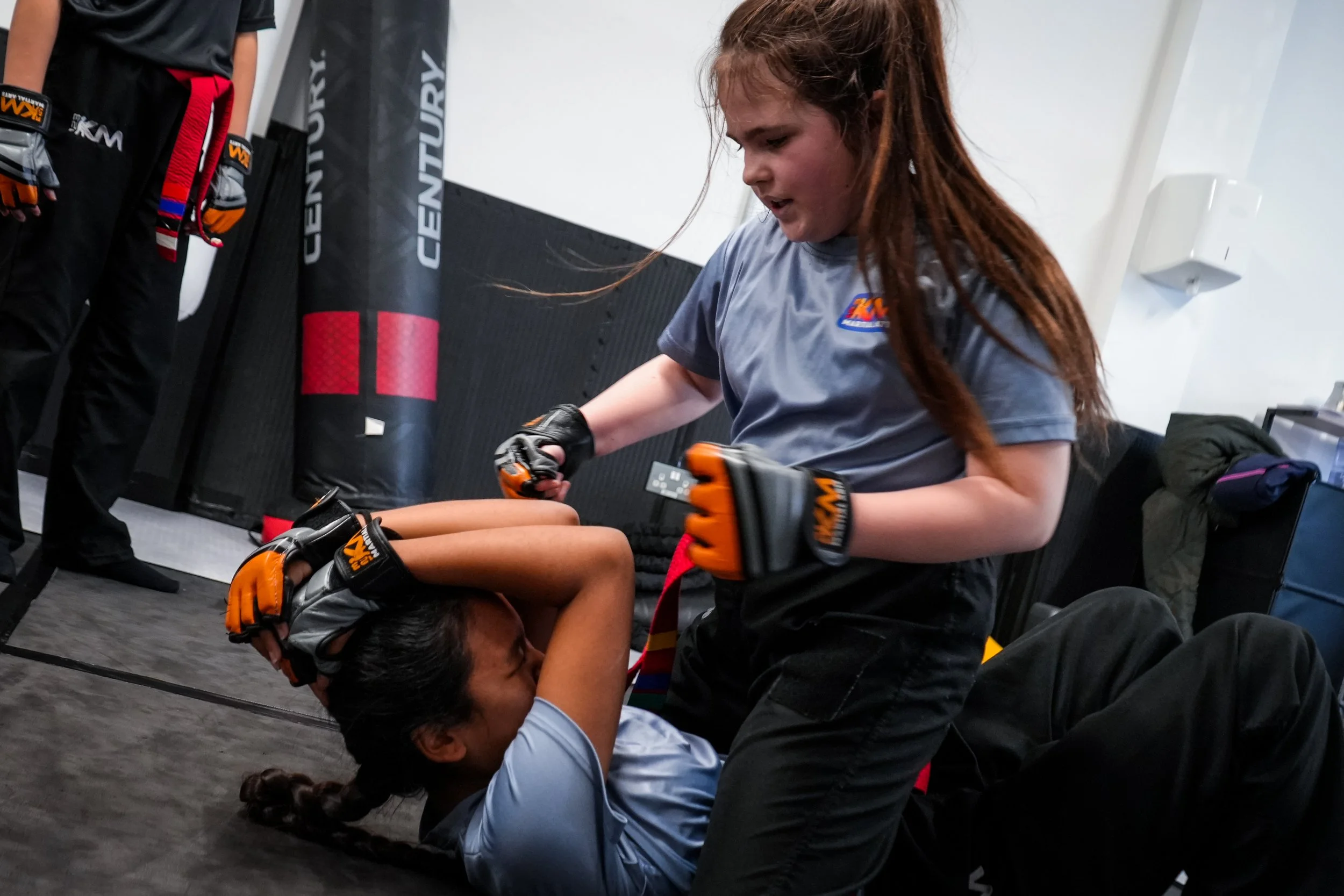 A girl with long red hair tied back, wearing a gray T-shirt, is practicing self-defense training with a partner in a martial arts gym. She is in a defensive position, blocking an attack from her partner, who is lying on the ground. The gym has a punching bag and black padded walls.